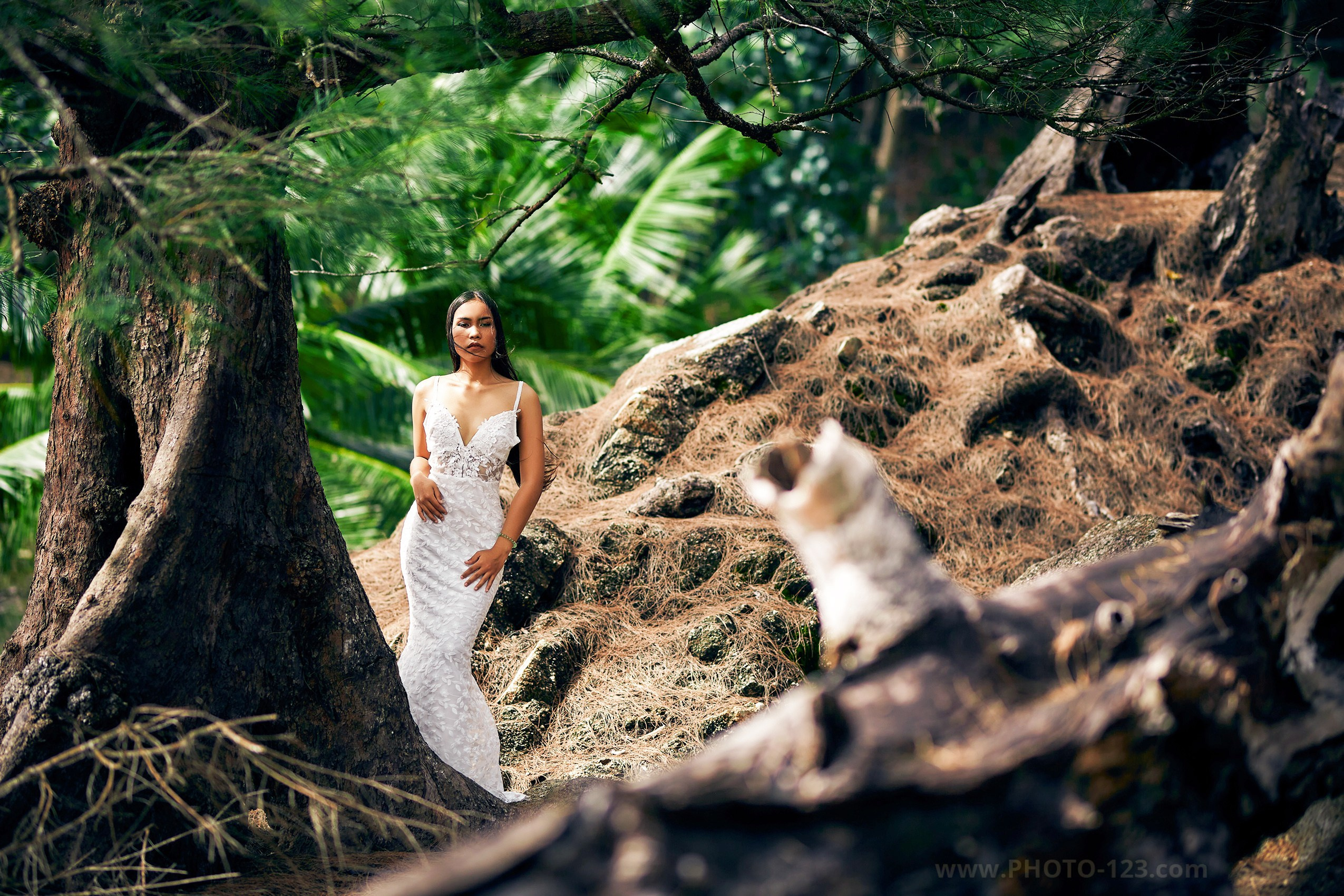 Woman in a white lace wedding dress standing among trees and rocks in a lush tropical forest, photographer in Phu Quoc