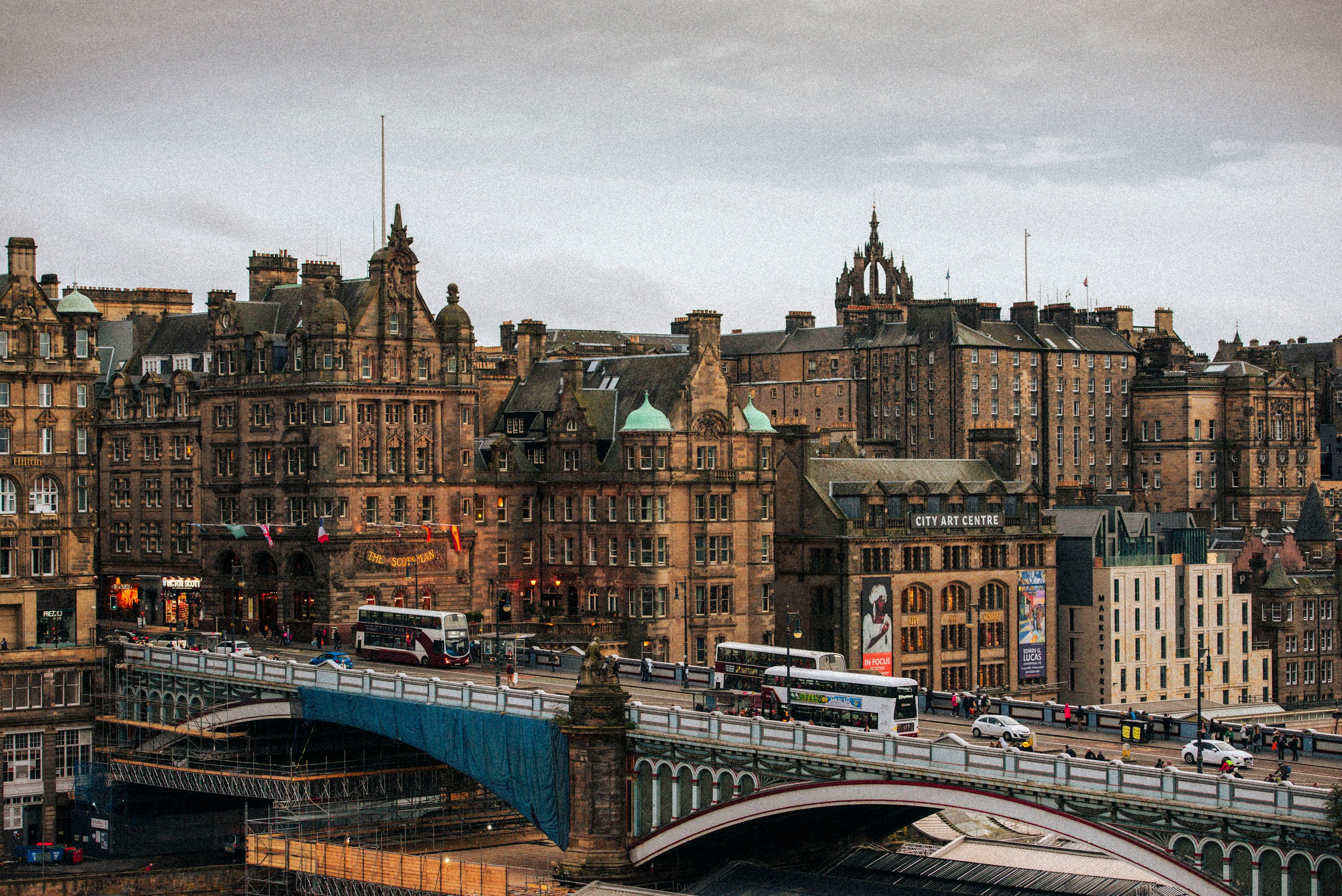 Панорамный вид на исторический Северный мост (North Bridge) в Эдинбурге, Шотландия, с красными двухэтажными автобусами и зданиями Старого города на заднем плане под пасмурным небом.
