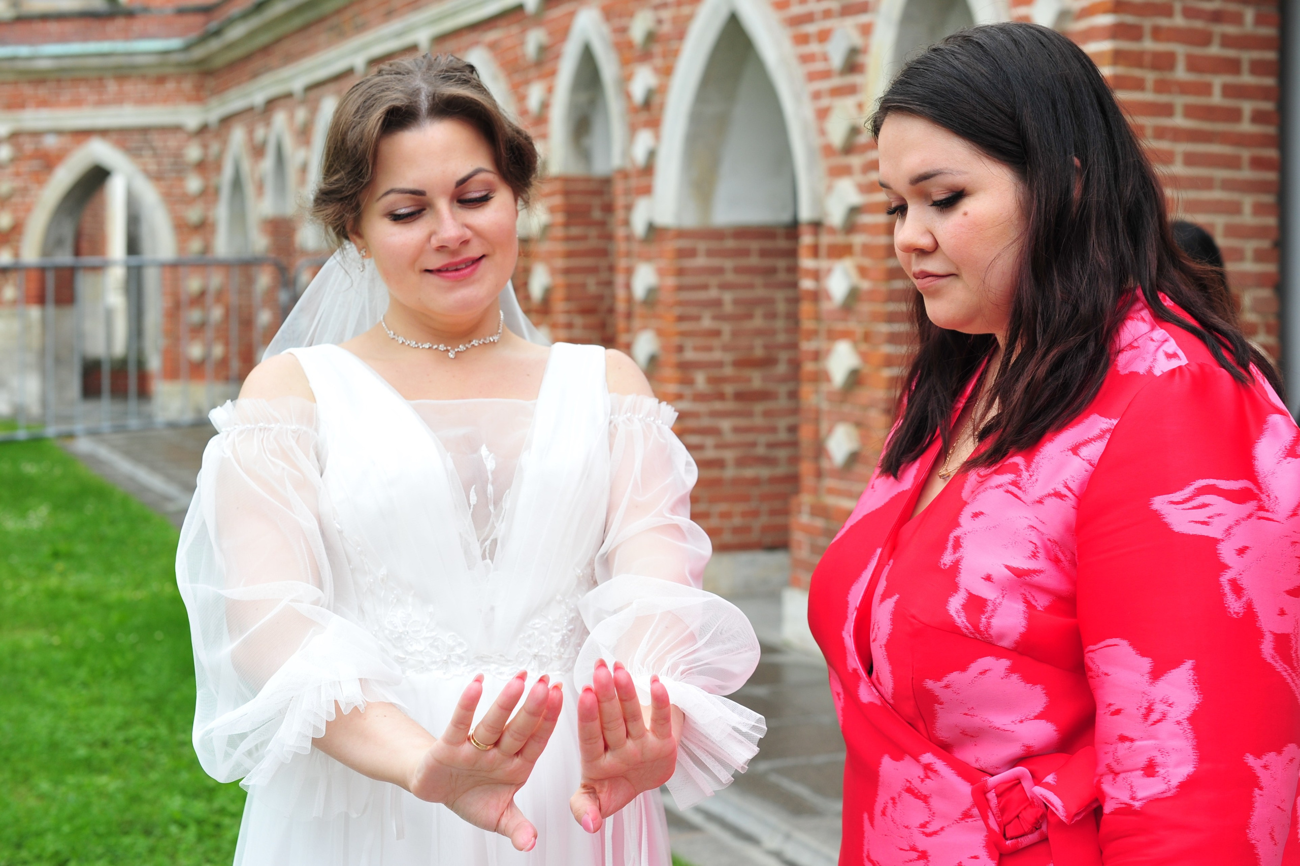 Свадьба в Царицыно пасмурным летним днем, невеста любуется обручальным кольцом и маникюром на своих руках, рядом стоит подруга невесты в красном кимоно 