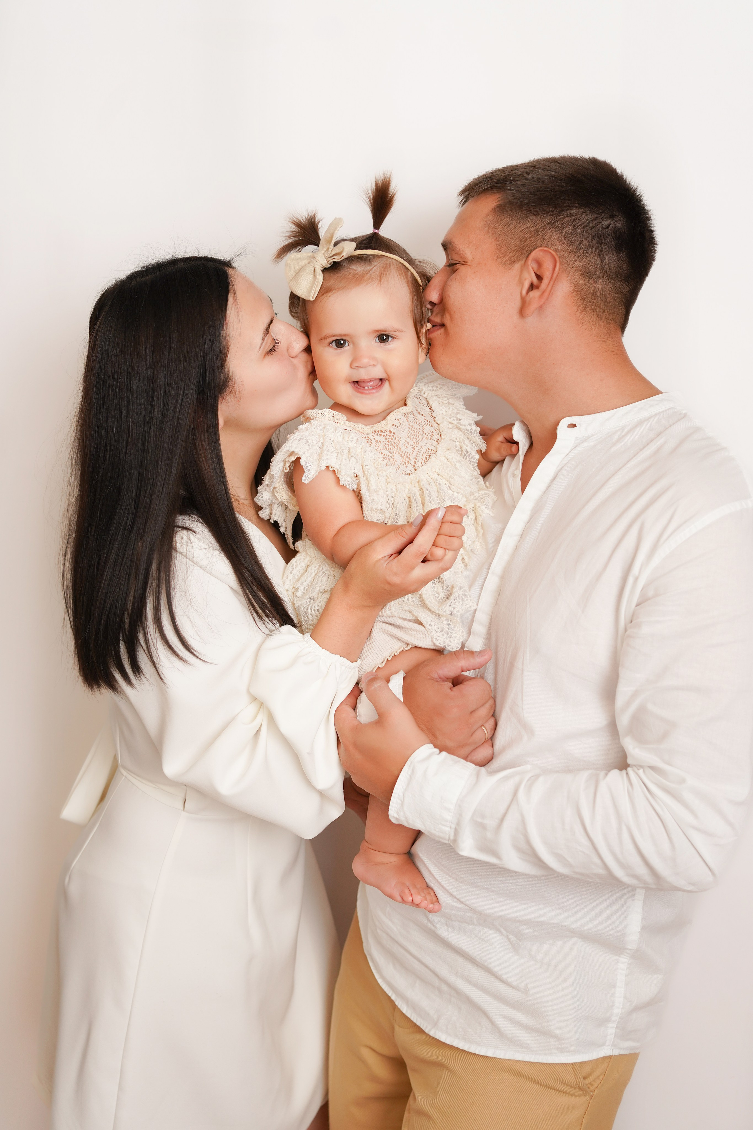 Семья приехала на фотосессию. На фото мама и папа целуют малышу. Она улыбается.