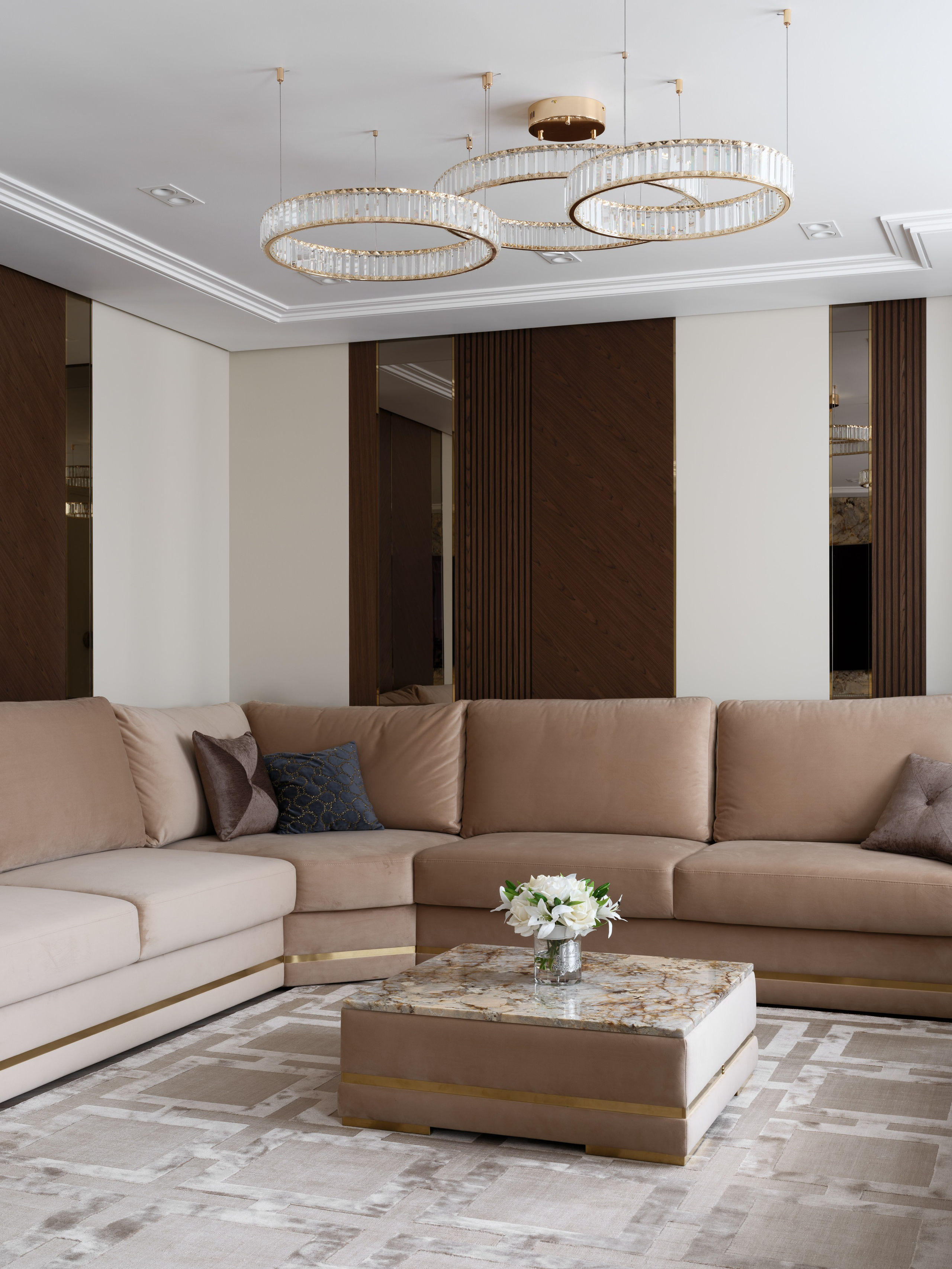 Просторная гостиная с большим угловым диваном в бежевой обивке, низким журнальным столиком из натурального камня и декоративными подушками, оформленная в спокойной нейтральной гамме с акцентом на современную люстру.