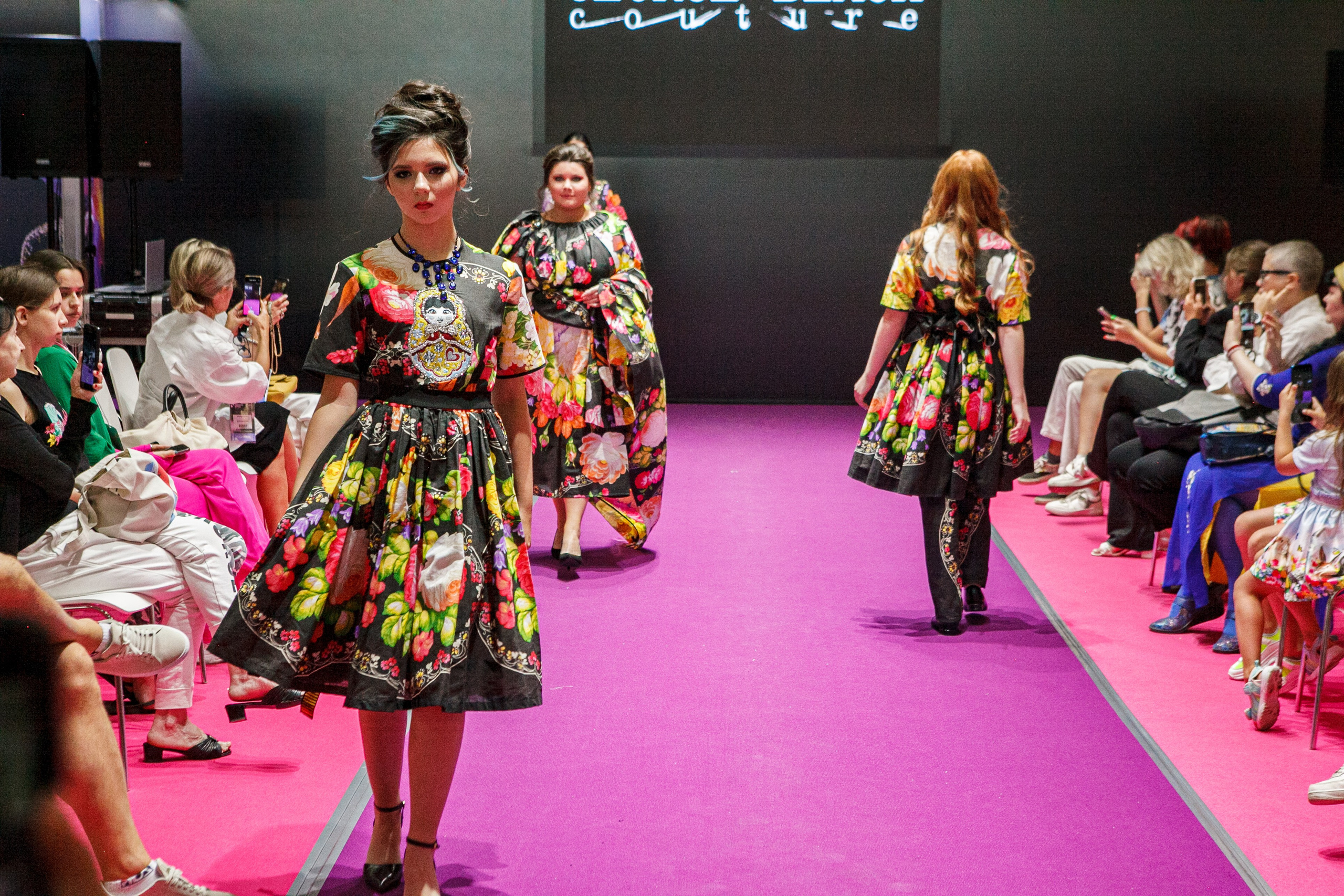  "Модели в ярких нарядах проходят по подиуму во время показа мод, освещенные яркими софитами и окруженные восторженными зрителями."