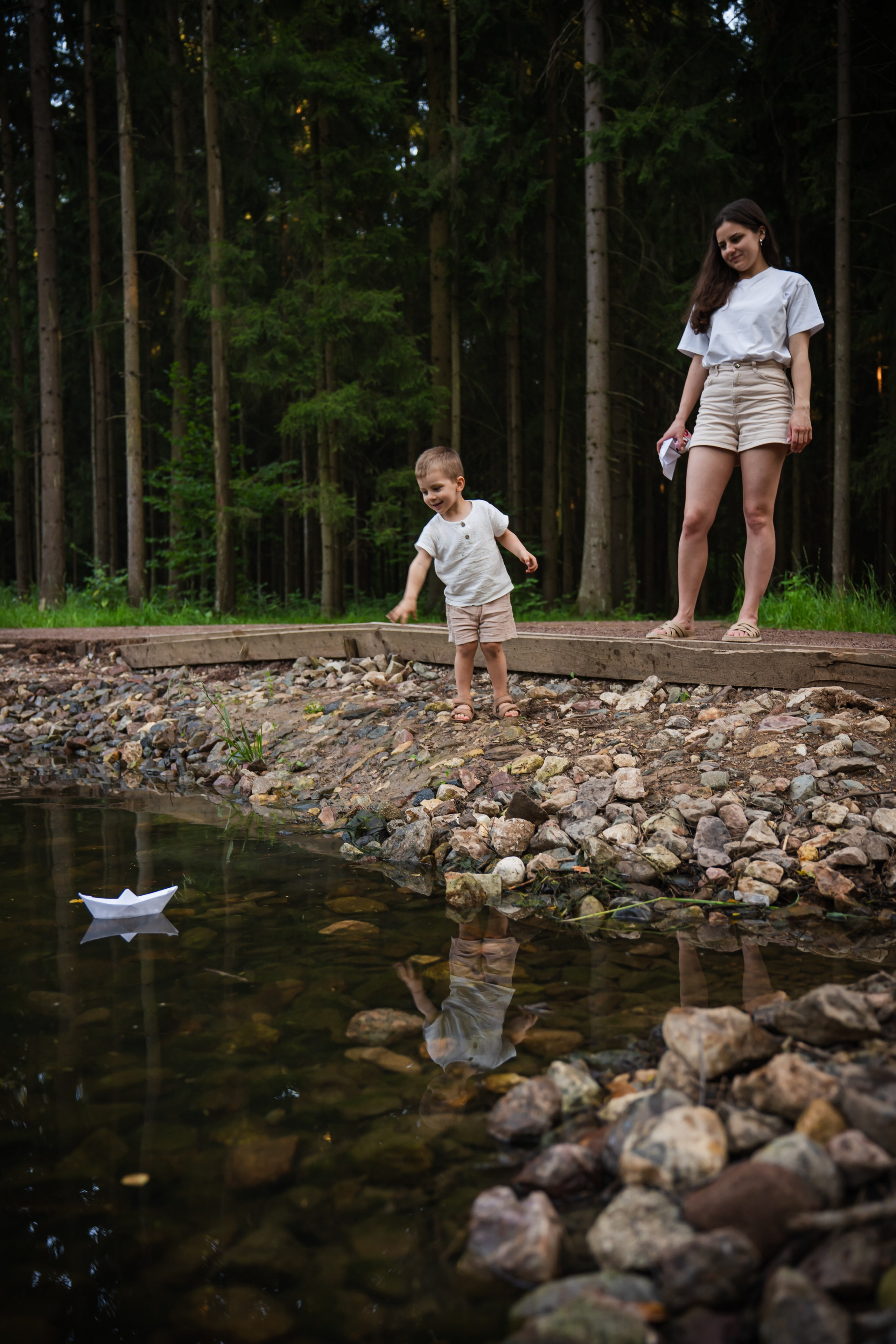 мама и сын в парке пускают бумажный кораблик в пруд