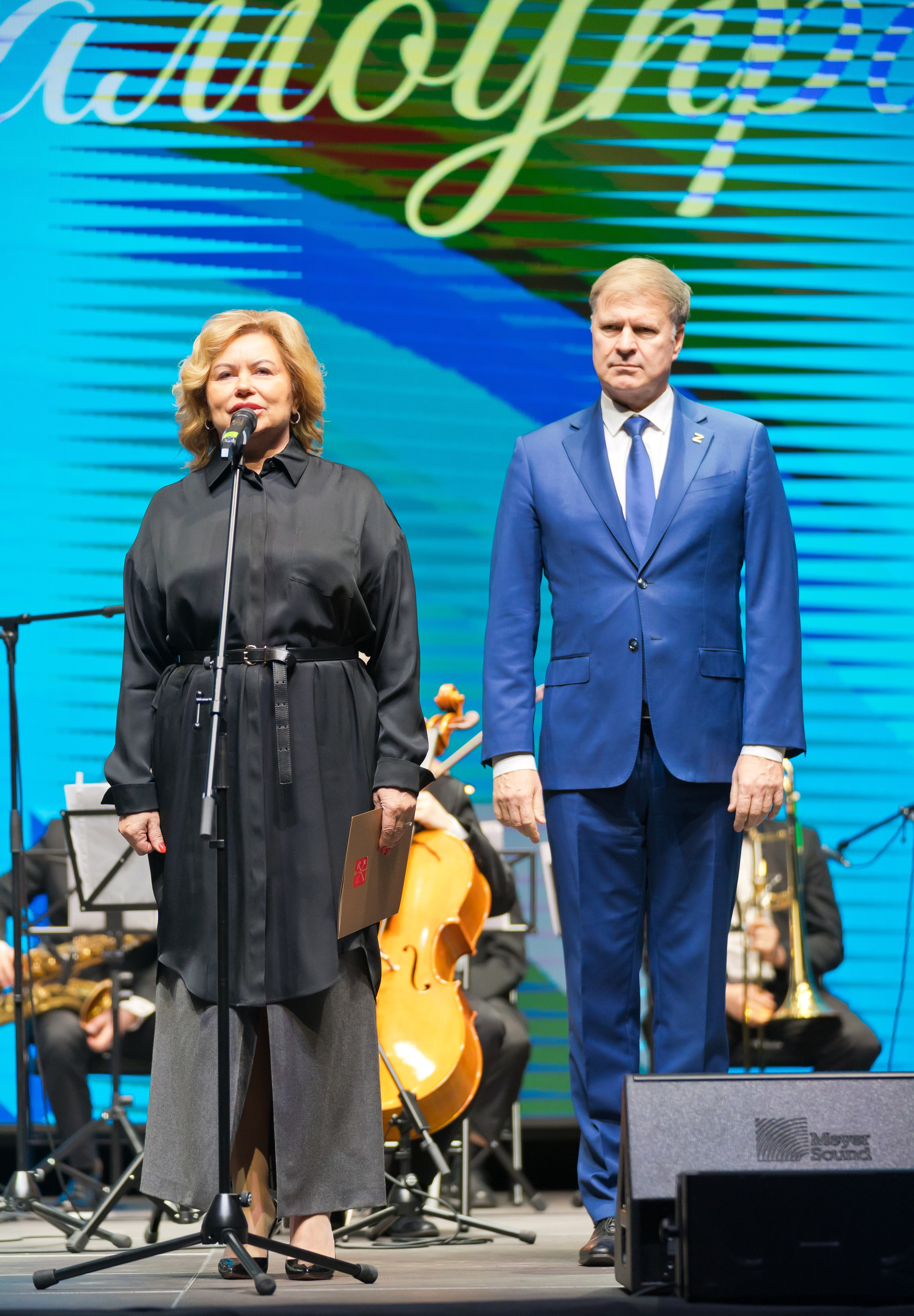 Женщина в чёрной блузке и серой юбке выступает с речью на сцене. Рядом стоит мужчина в синем костюме. День местного самоуправления, Санкт-Петербург. Фото Евгения Мальцева.