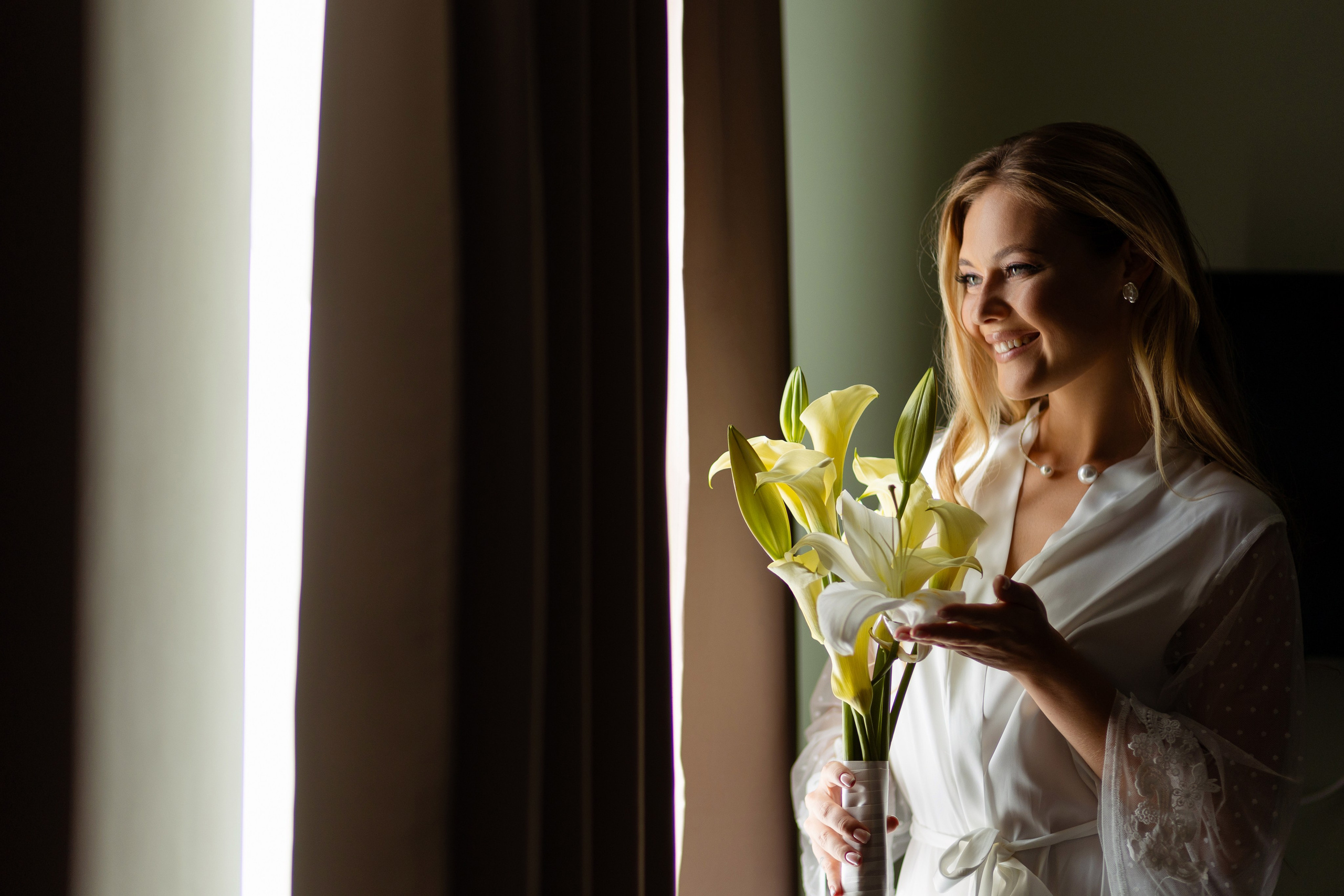 Невеста стоит у окна с букетом желтых цветов.