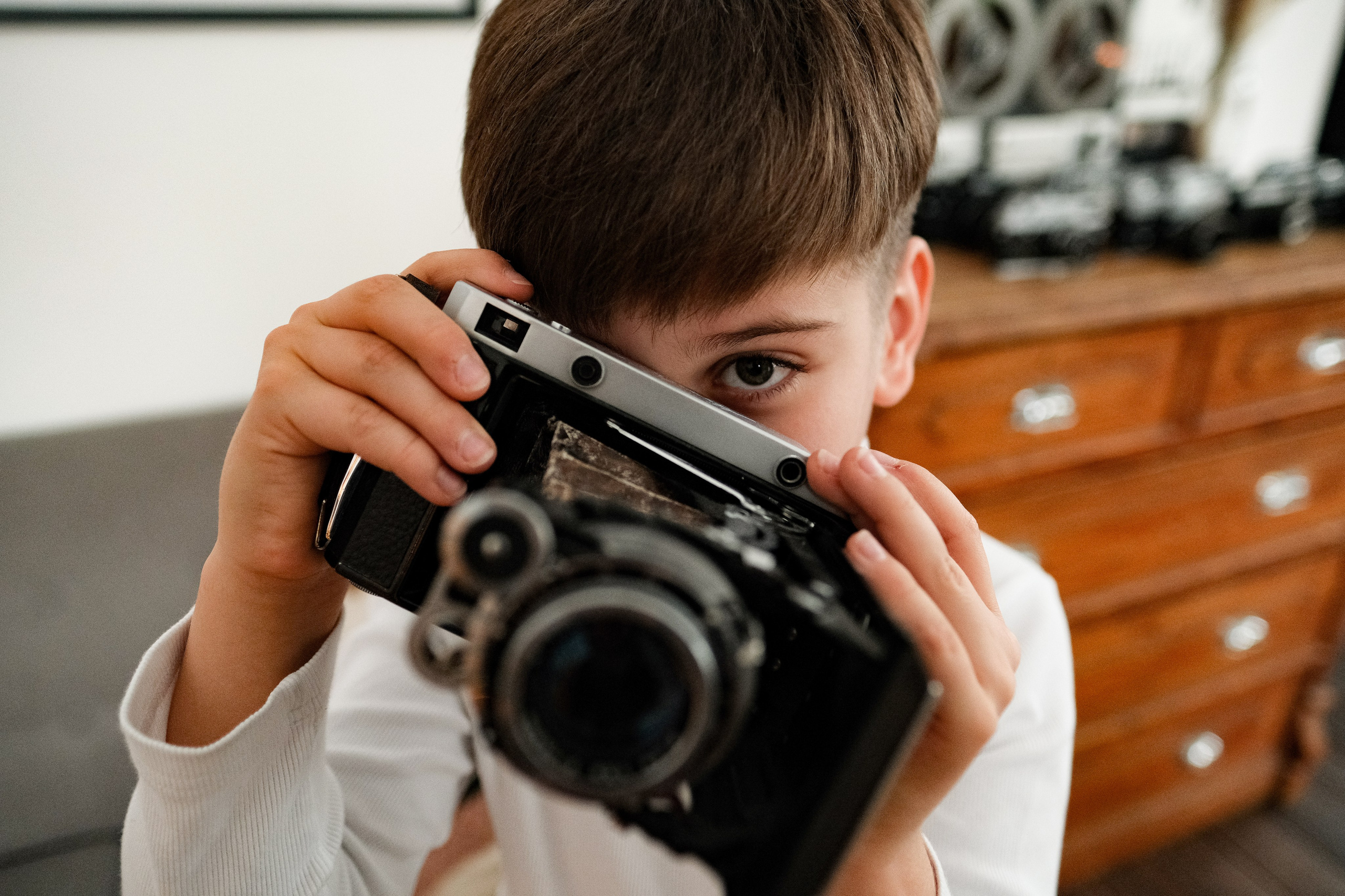 Мальчик с хитрым взглядом выглядывает из-за старого фотоаппарата. Мальчик с фотоаппаратом.