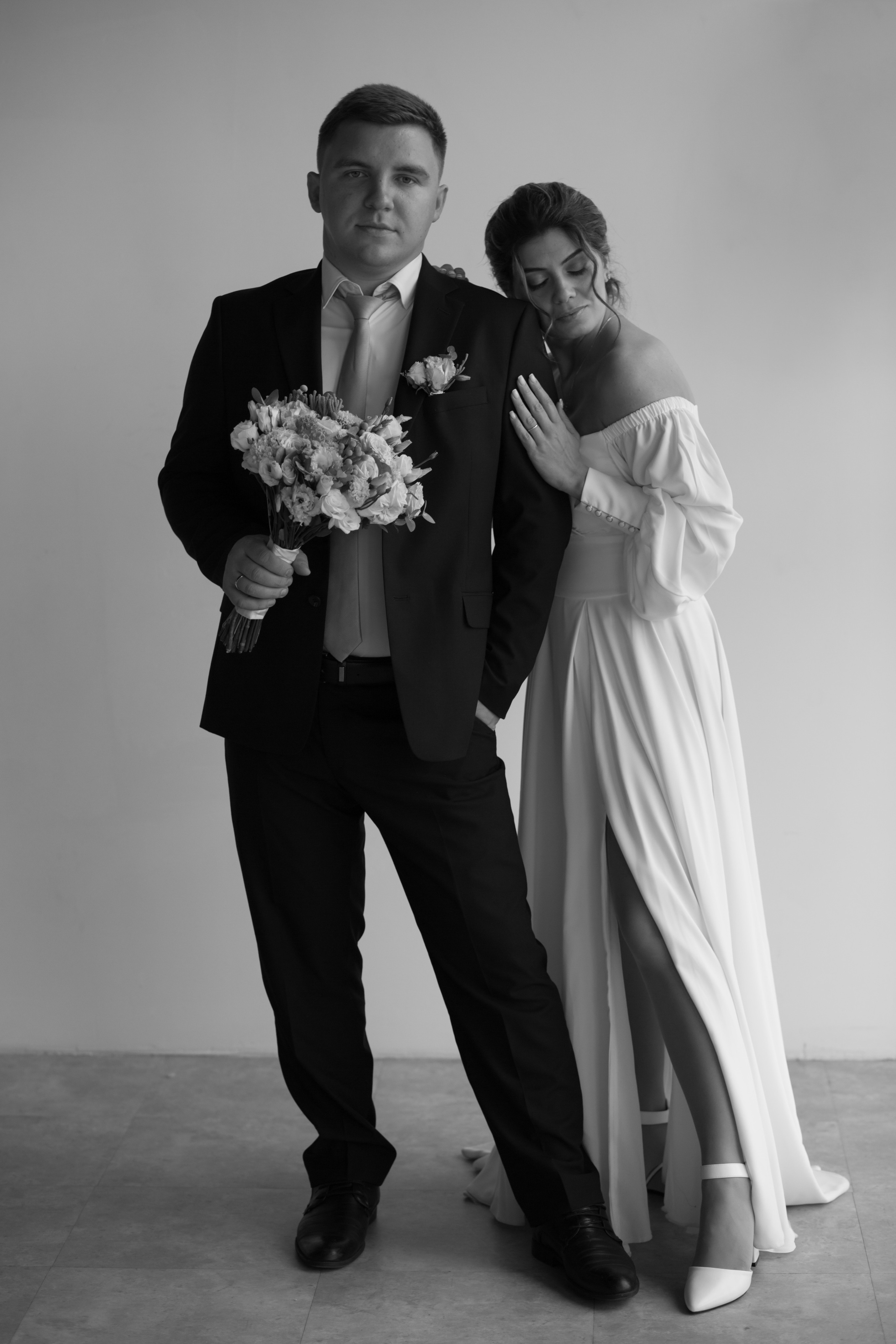 у жениха в руках букет цветов на фоне белой стены рядом стоит невеста в белом платье с разрезом