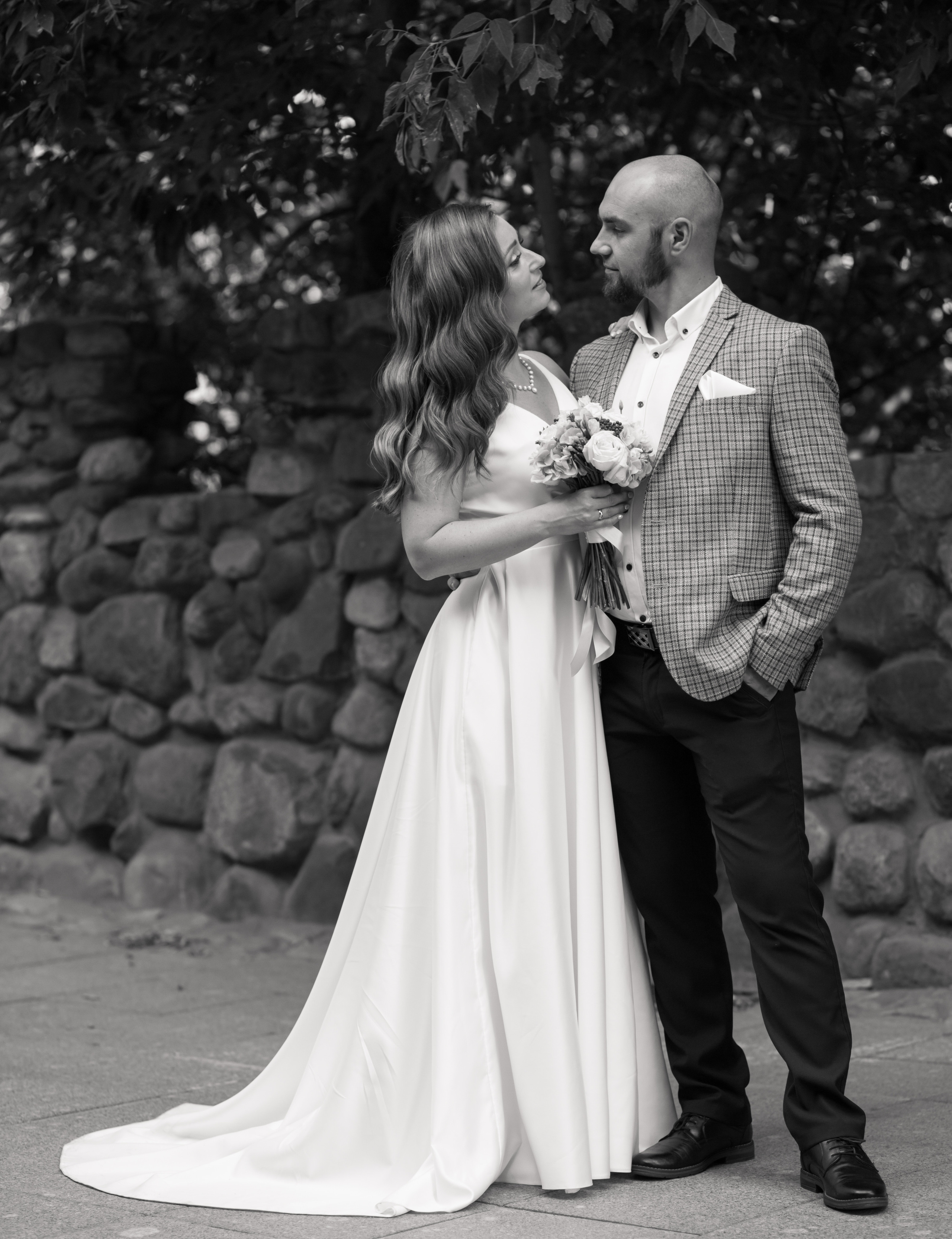 у невесты в руках букет цветов, жених обнимает ее за талию, смотрят нежно друг-другу в глаза