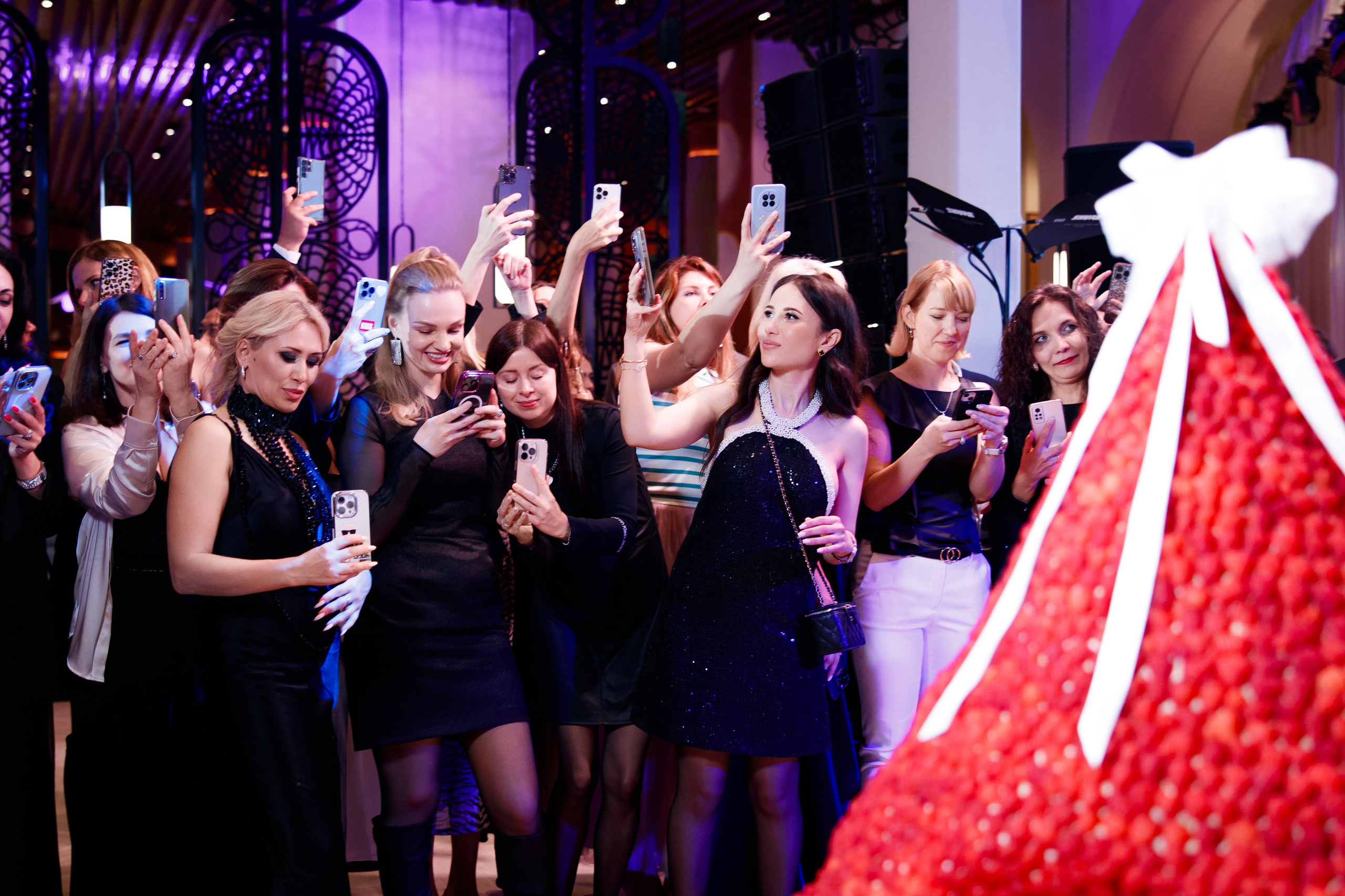 Группа гостей в вечерних образах увлеченно снимает на смартфоны яркую инсталляцию — огромную клубничную пирамиду с белым бантом. Атмосфера современного светского события.