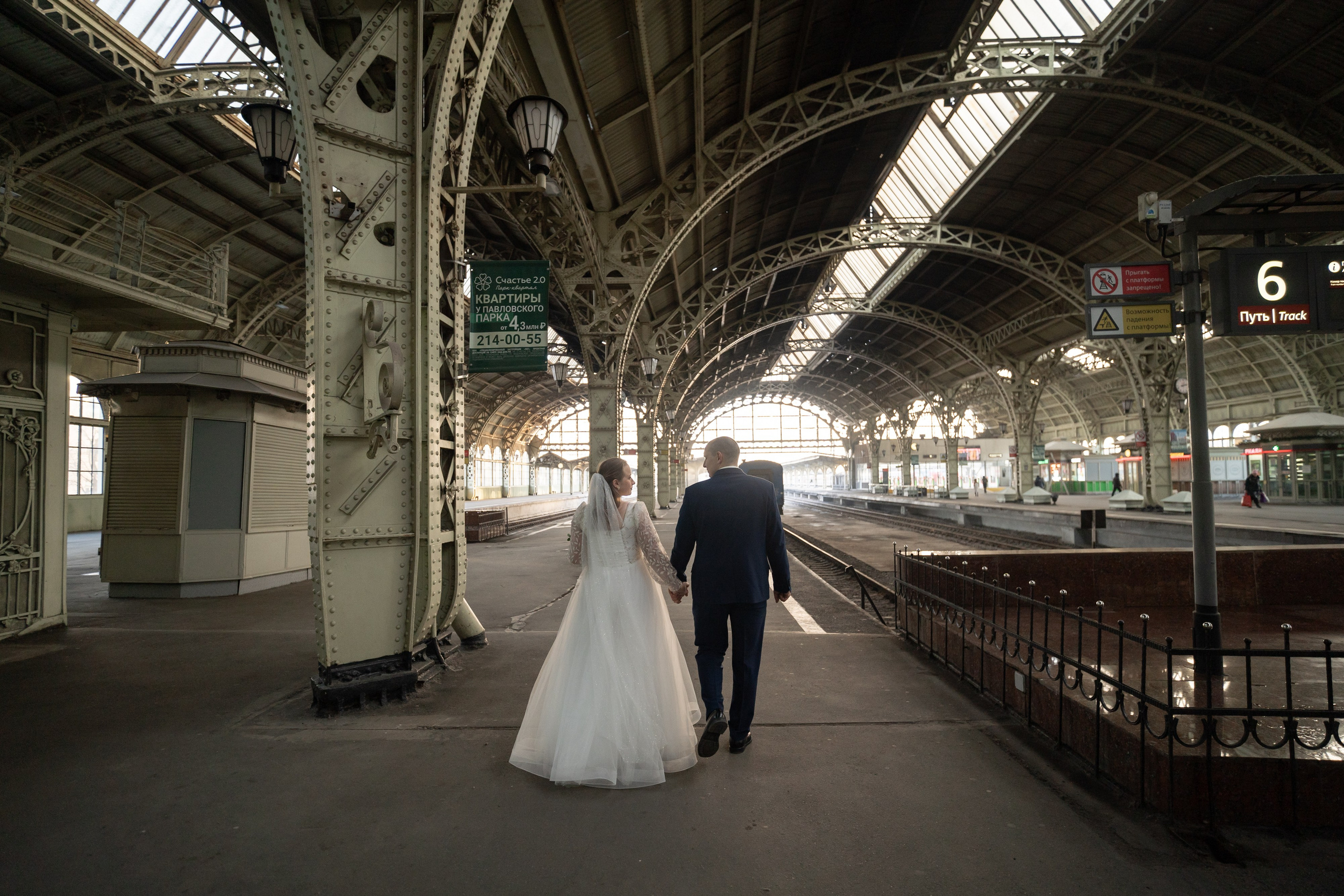 Свадебная фотография на Витебском вокзале в Санкт-Петербурге Жених и невеста за руки выходят на перрон под полукруглыми сводами.