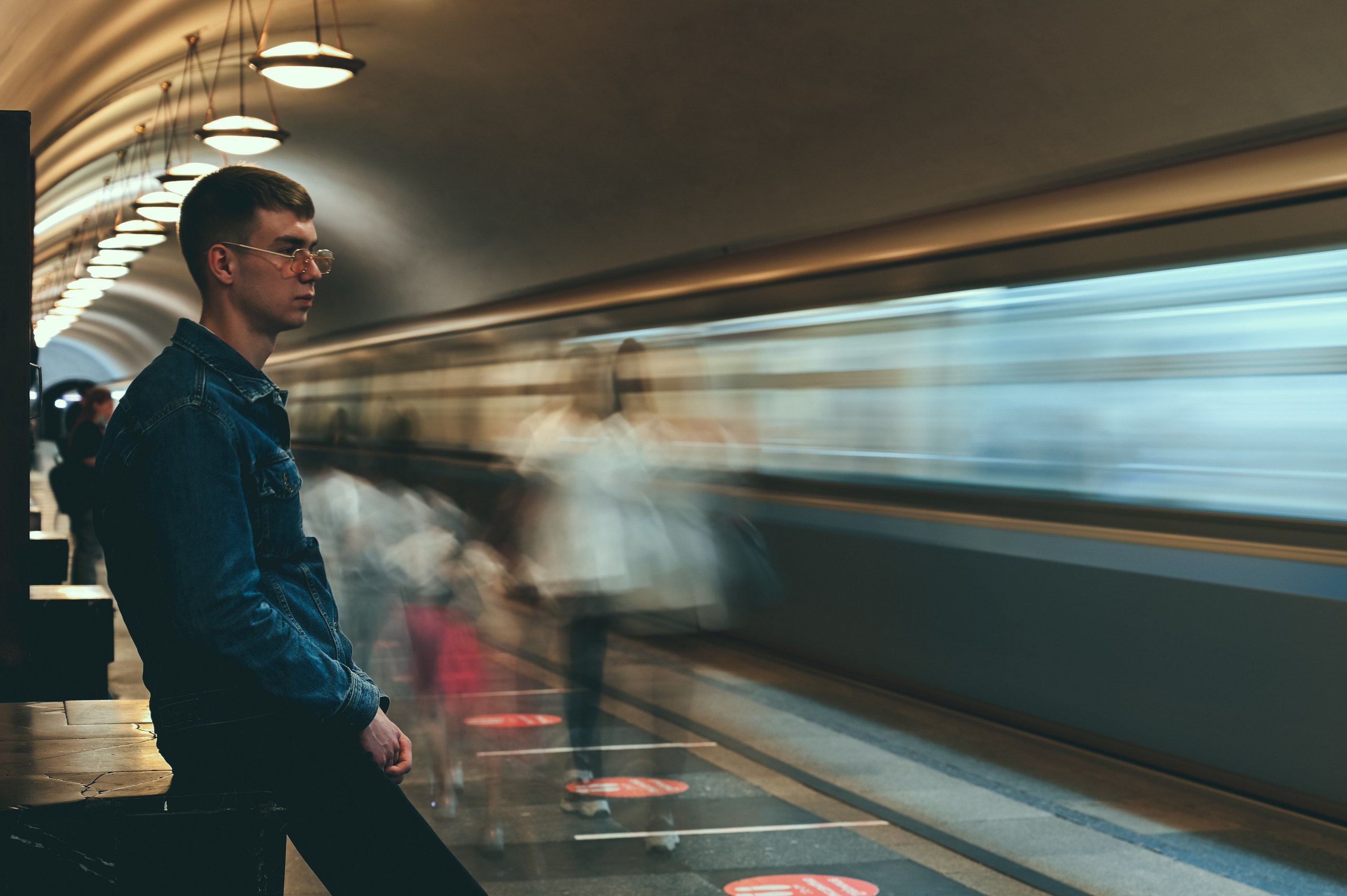 Молодой человек в джинсовом пиджаке стоит на платформе станции метро, глядя в сторону. На заднем плане виден поезд, который движется мимо него, создавая эффект размытия. Освещение станции подчеркивает современный дизайн интерьера с подвесными светильниками и металлическими стенами. Вокруг модели несколько других людей, также движущихся или стоящих, что добавляет динамичность к сцене. Фотография передает атмосферу ожидания и спокойствия в условиях городской среды.