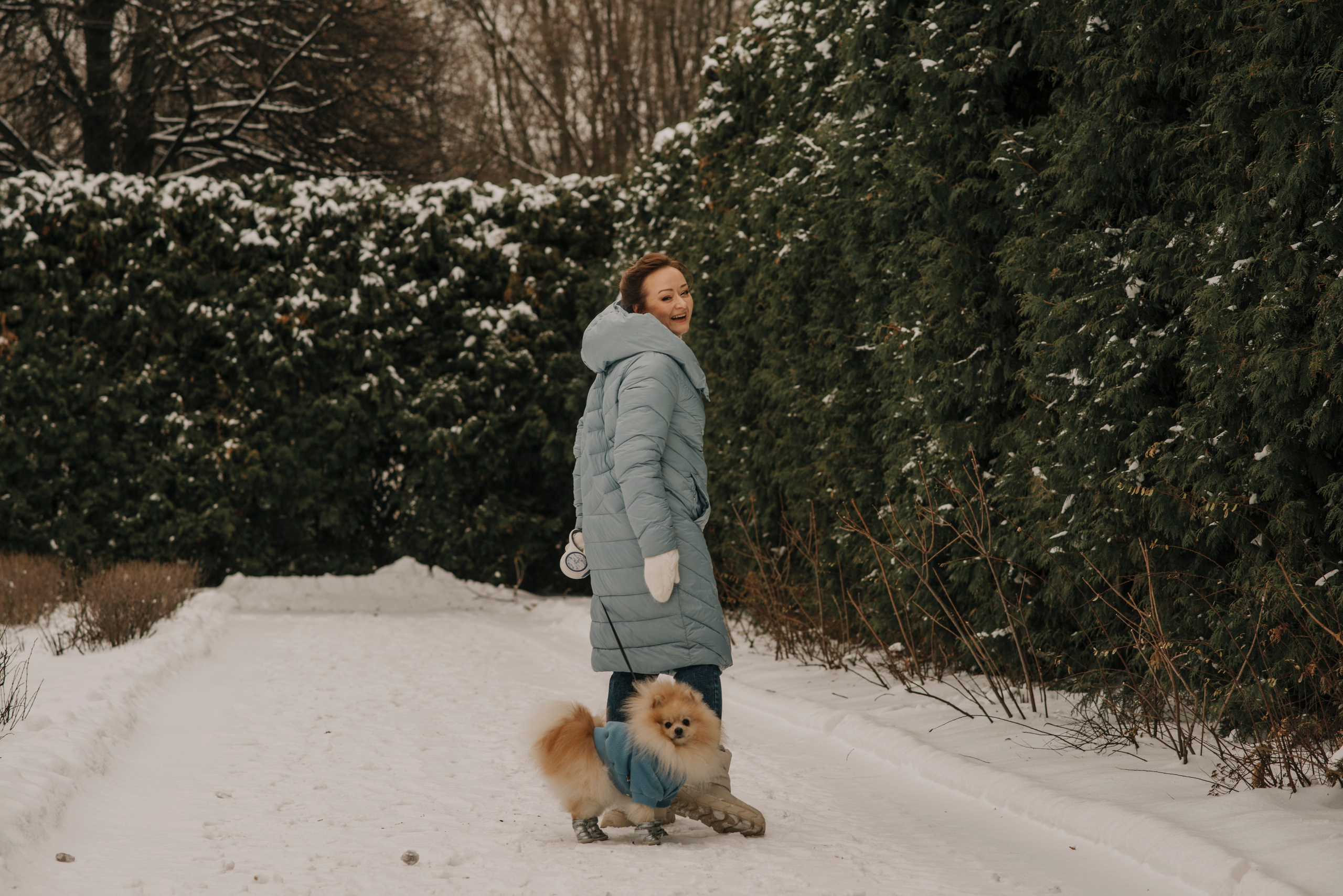 Женщина в голубом пуховике гуляет с собакой шпицем по зимнему парку