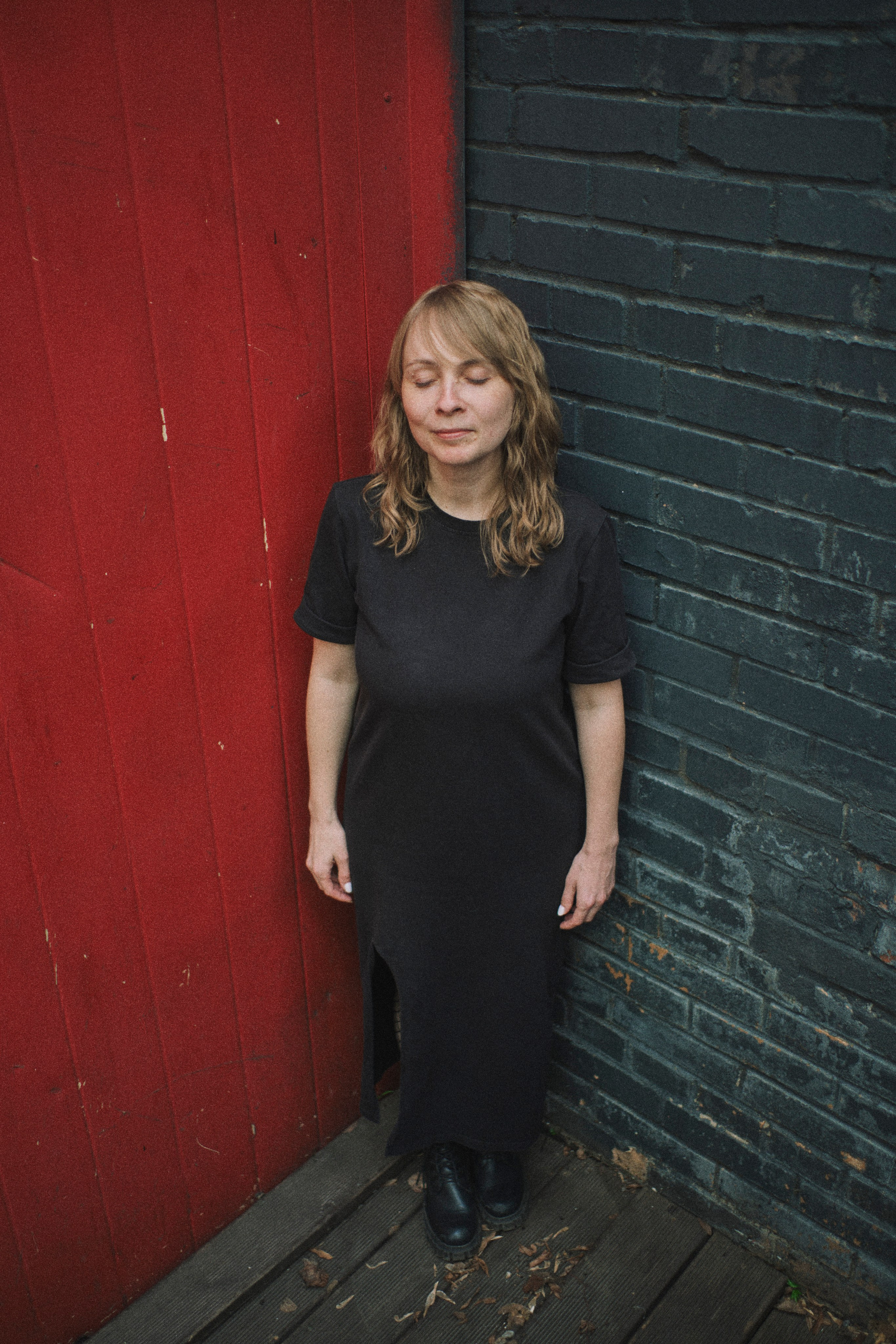 Женщина стоит в углу между красной и черной стеной, закрыв глаза