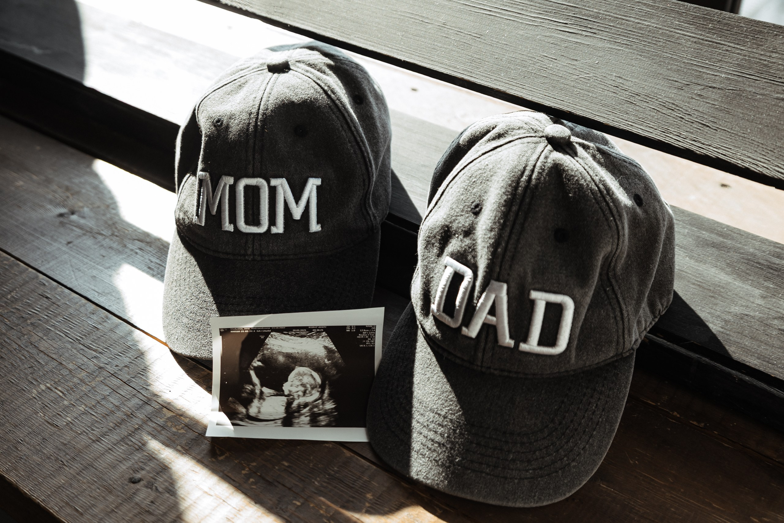 Предметный кадр: две бейсболки с надписями "MOM" и "DAD" рядом со снимком УЗИ, символ ожидания ребёнка.  