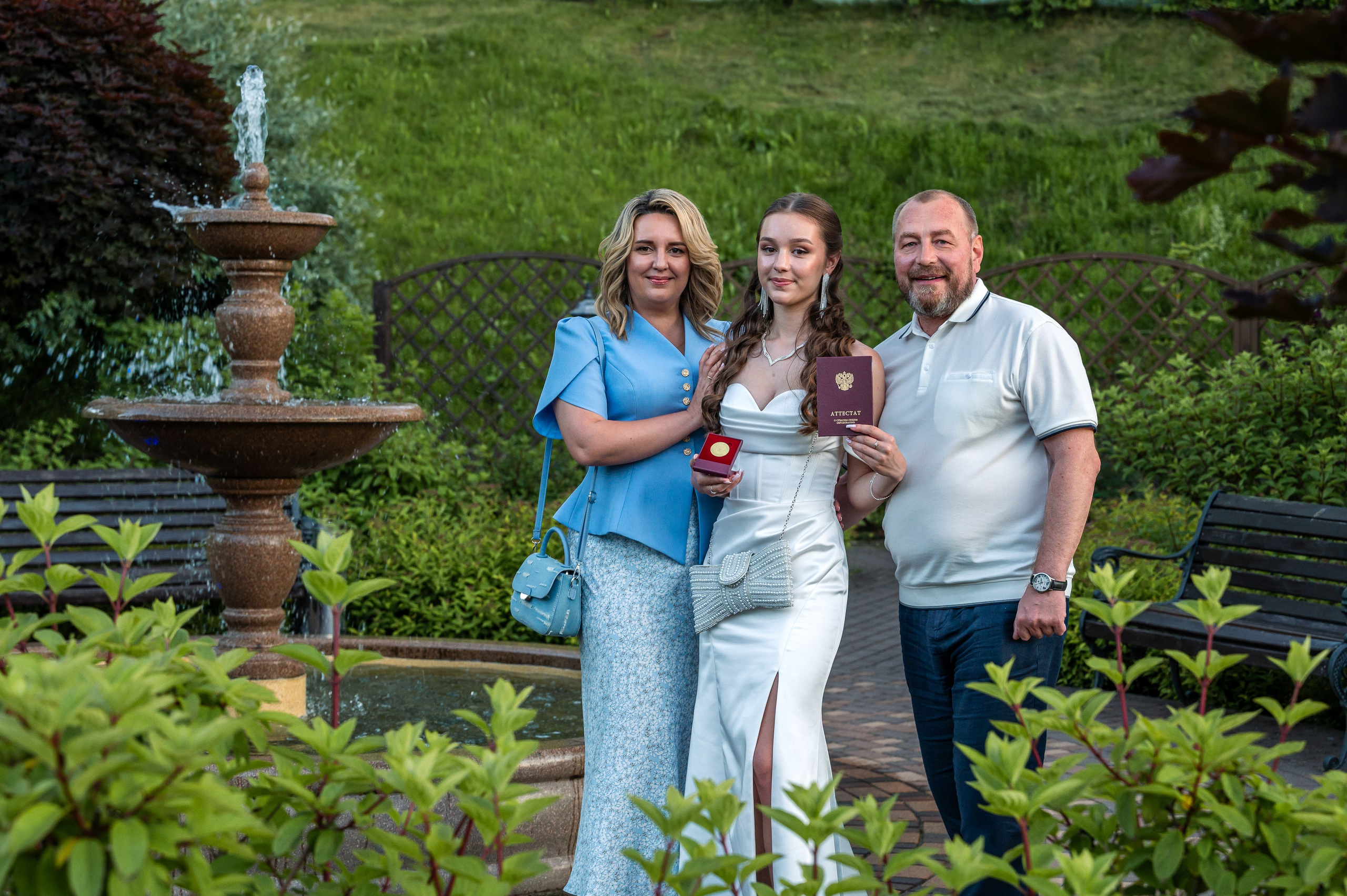 Выпускница-обладательница золотой медали вместе с родителями позируют фотографу в парковой зоне ресторана Городок