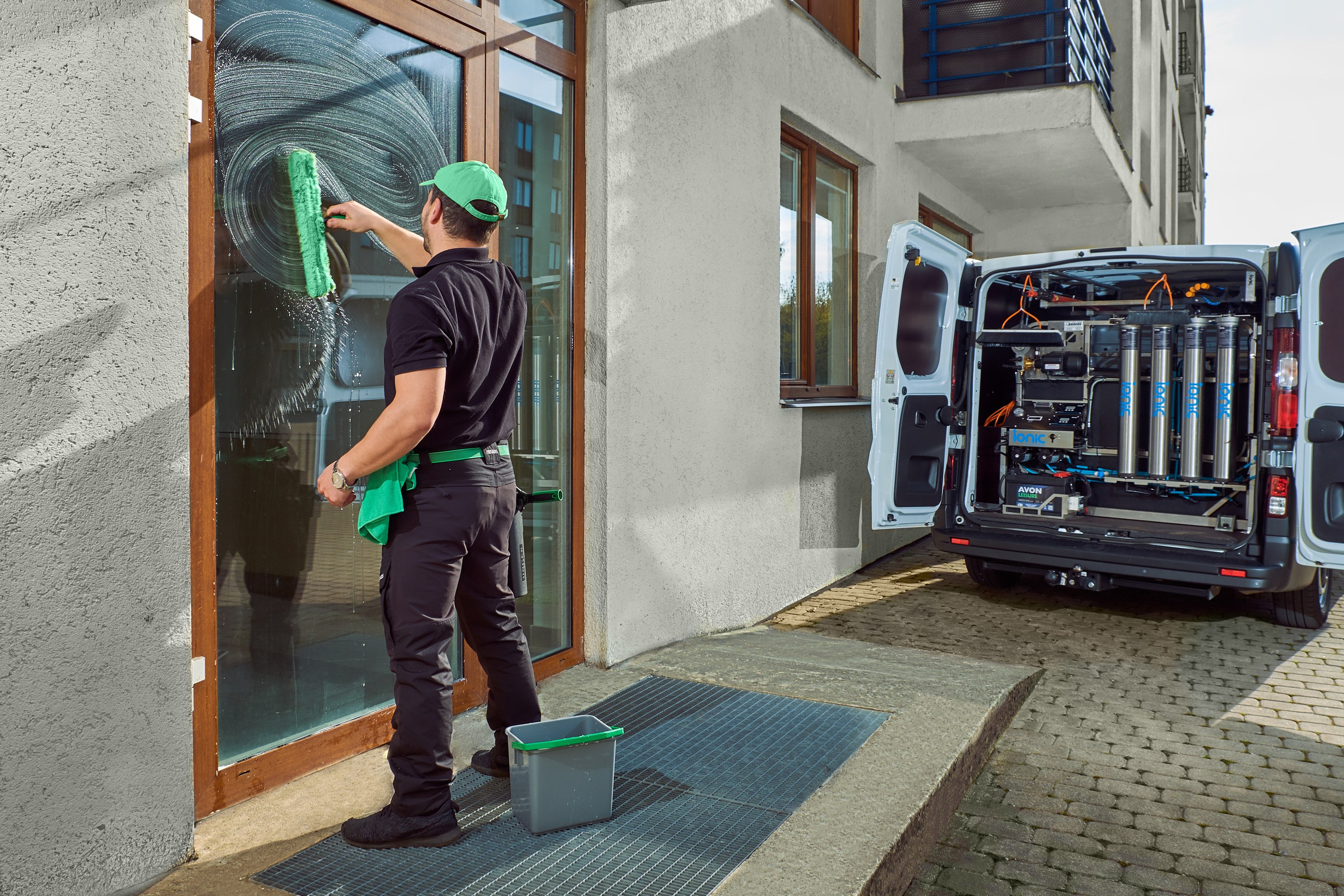 Профессиональная съёмка услуги по мойке витражных окон: специалист клининговой компании работает у входа в здание, рядом — открытый фургон с оборудованием. Контент для корпоративного сайта