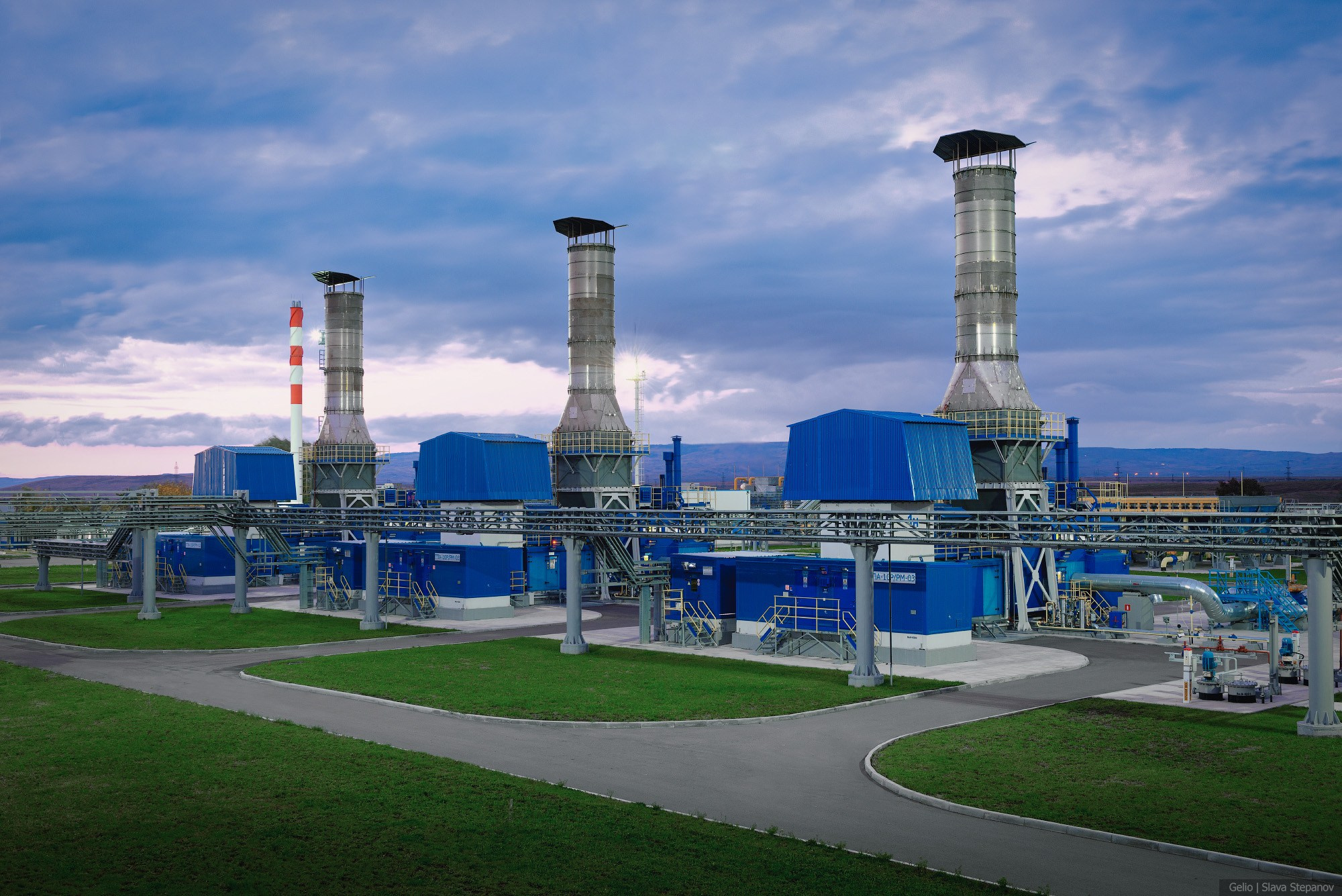 Современная газокомпрессорная станция с синими корпусами и высокими выхлопными трубами, чистая и высокотехнологичная территория промышленного объекта газовой отрасли, Gelio.