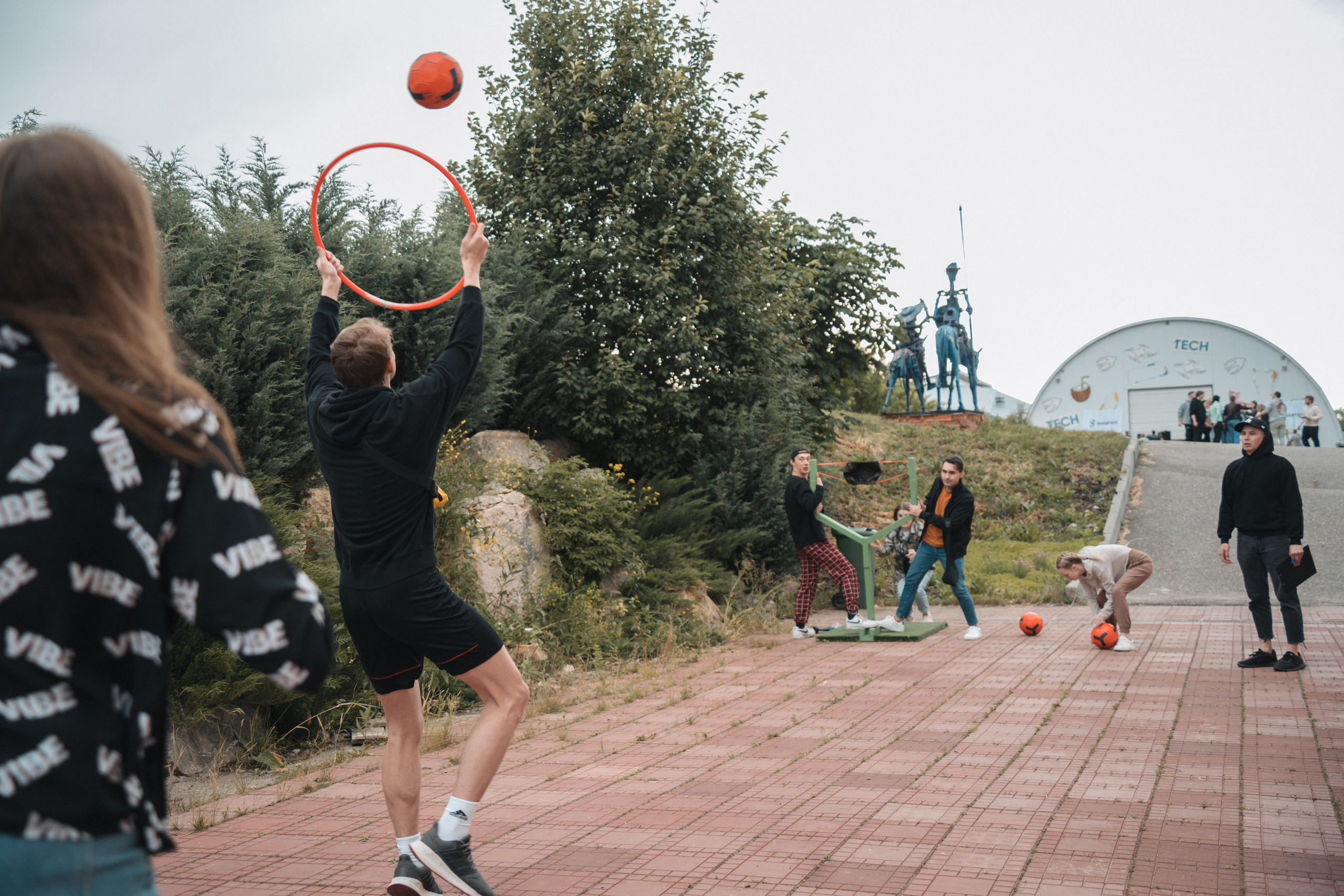 Молодые люди играют с оранжевым мячом и обручем на улице, вокруг несколько человек, летний день