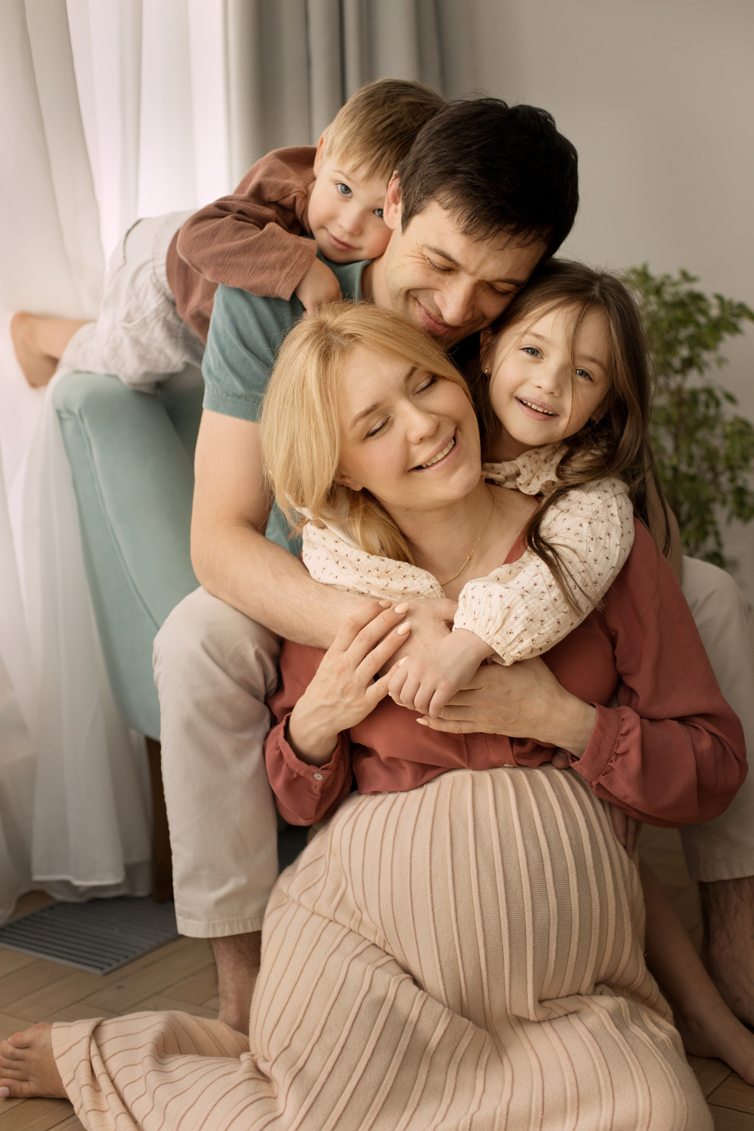 Беременная мама, папа, дочка и сын обнимаются сидя у окна.
