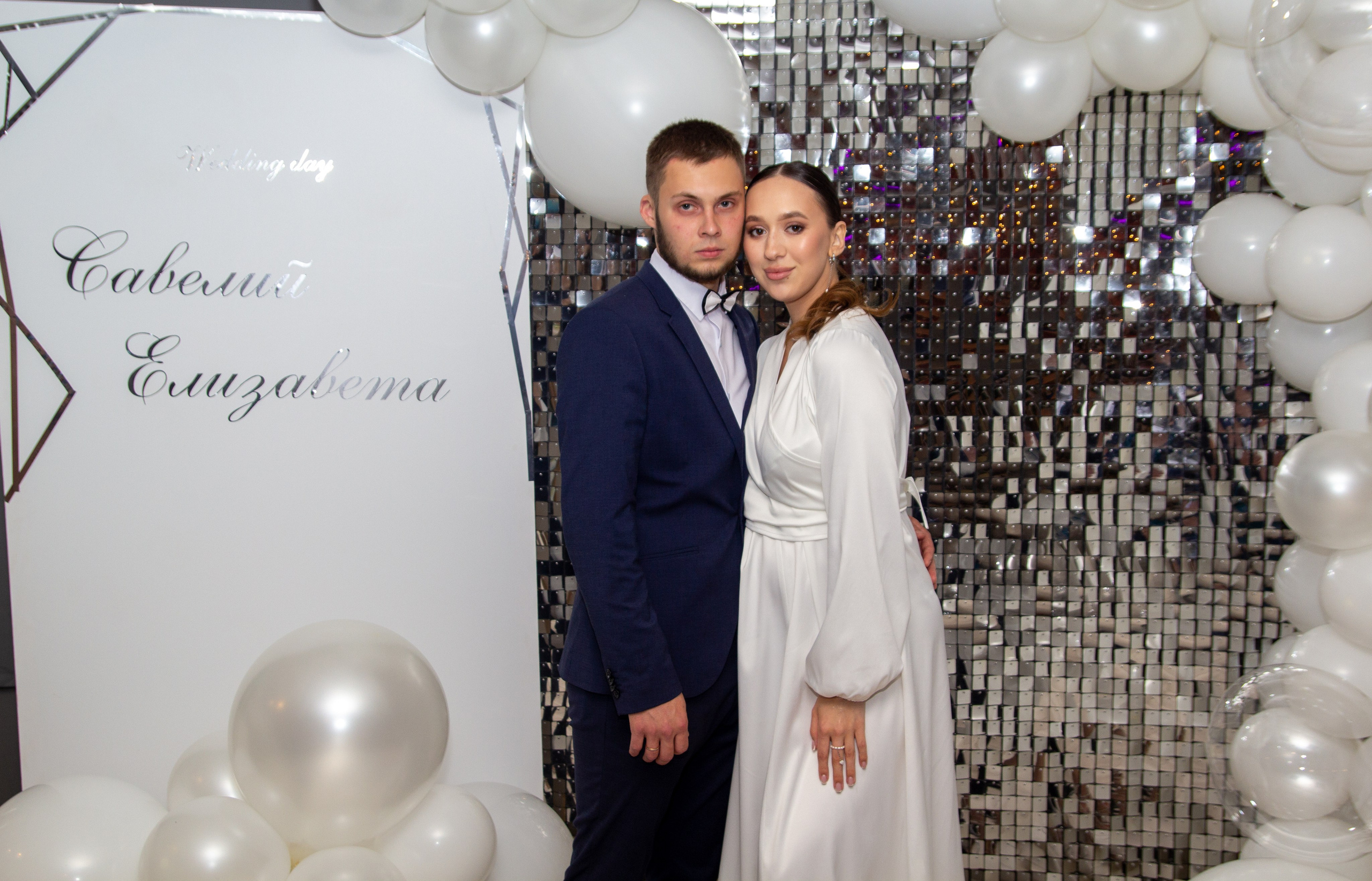 Пара позирует на фоне белого свадебного фотозоны с шарами и надписью.