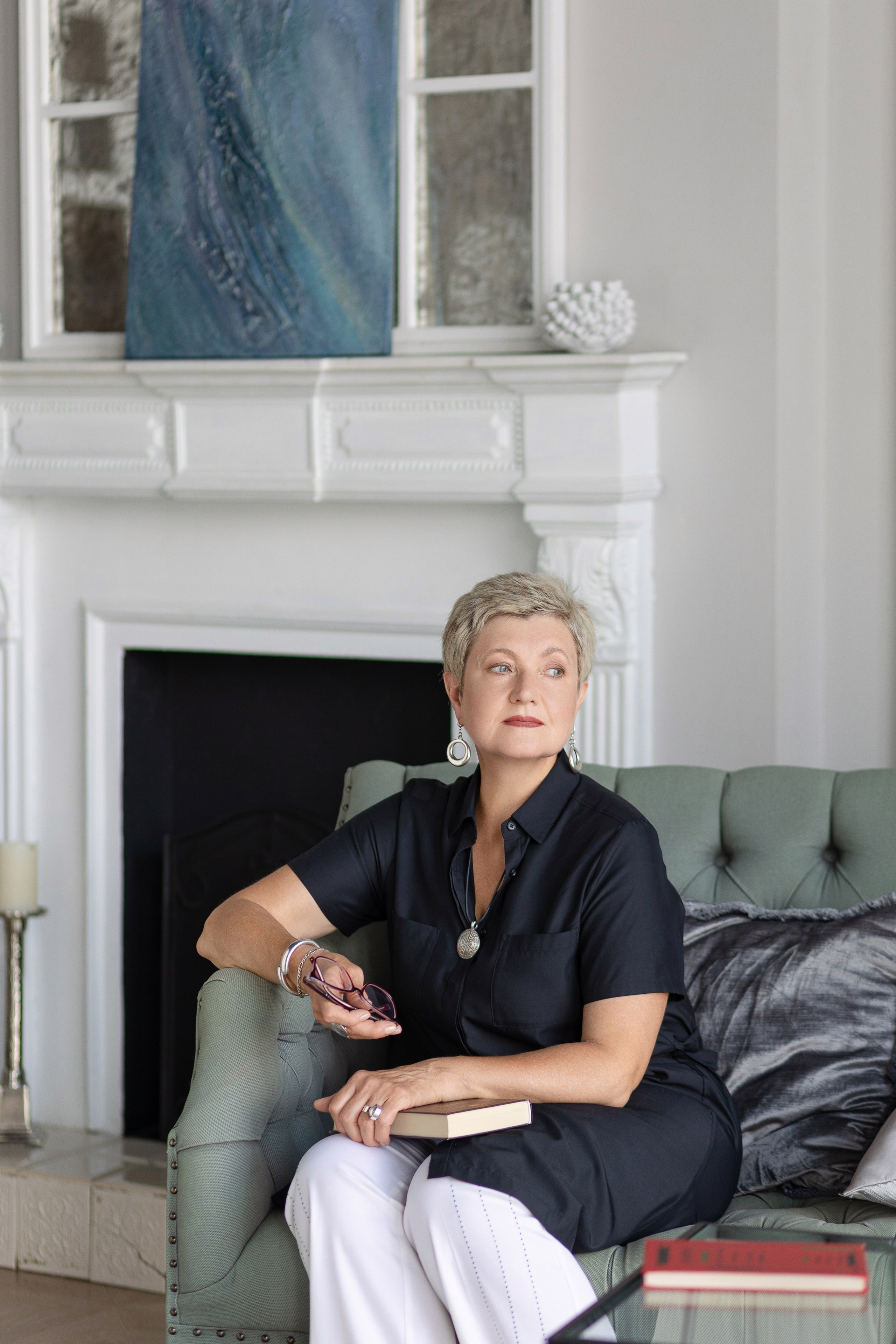 Женщина сидит на диване, положив руку на подлокотник, слева на диване лежит подушка.