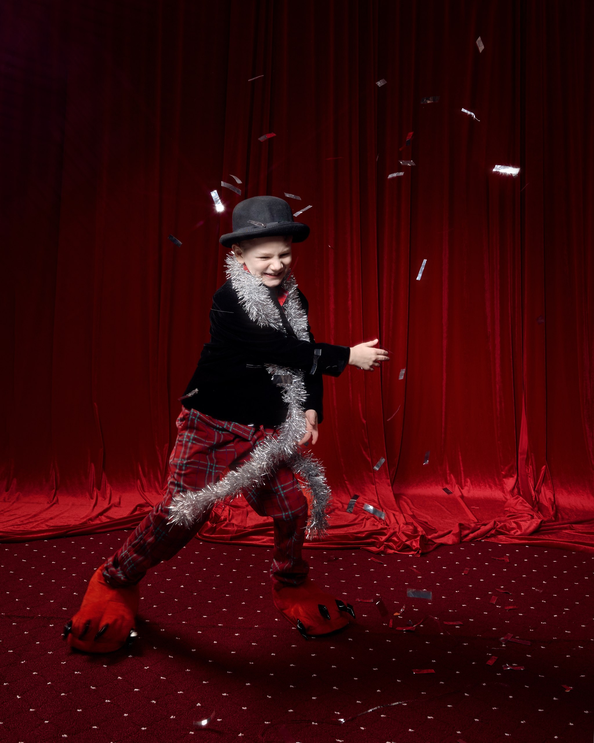 Динамичная детская фотосессия: мальчик в пижаме и шляпе танцует на красной сцене среди летящего конфетти. Семейный фотограф ловит момент радости и энергии ребенка. 