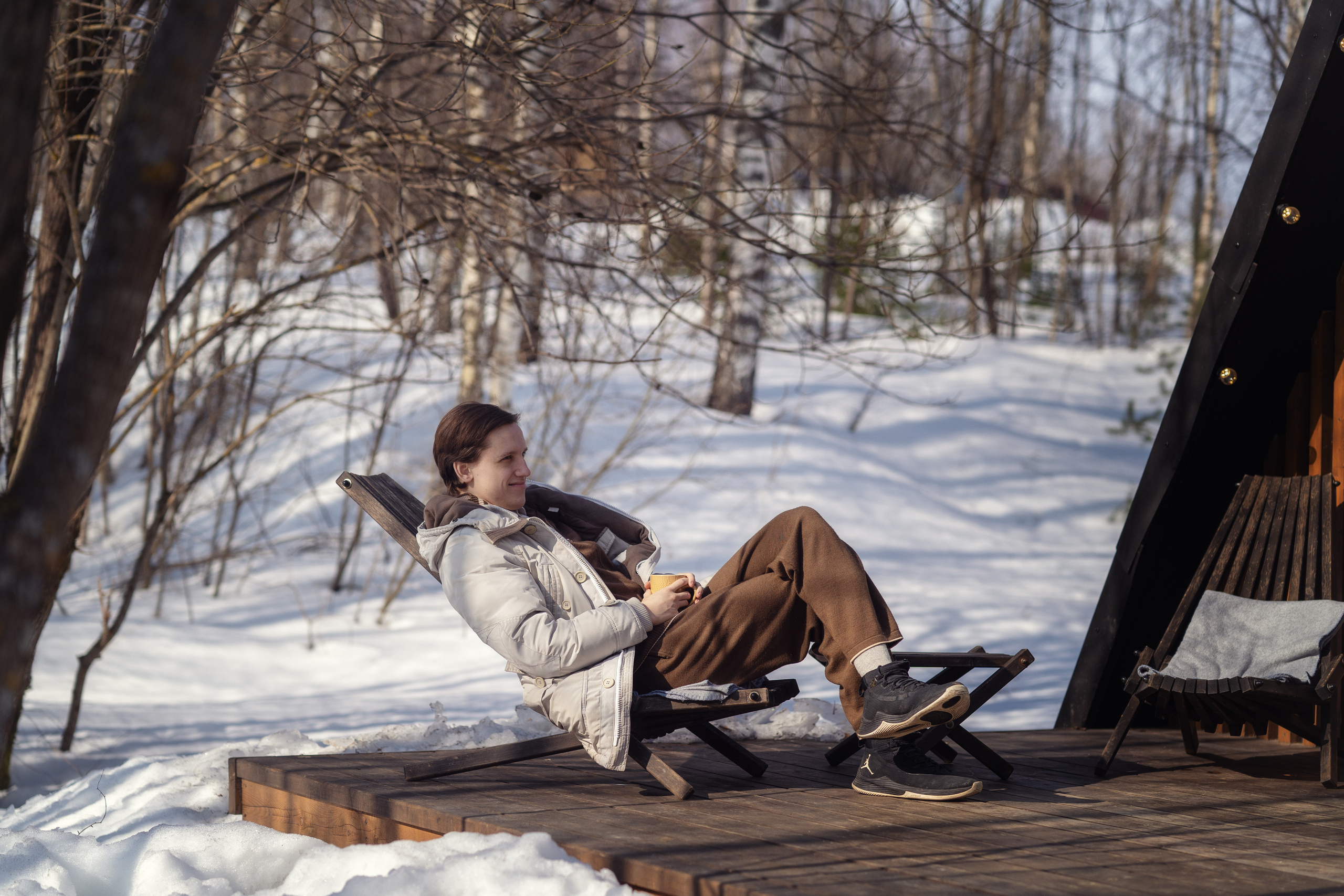 Молодой человек сидит в шезлонге на террасе A-frame домика в глэмпинге