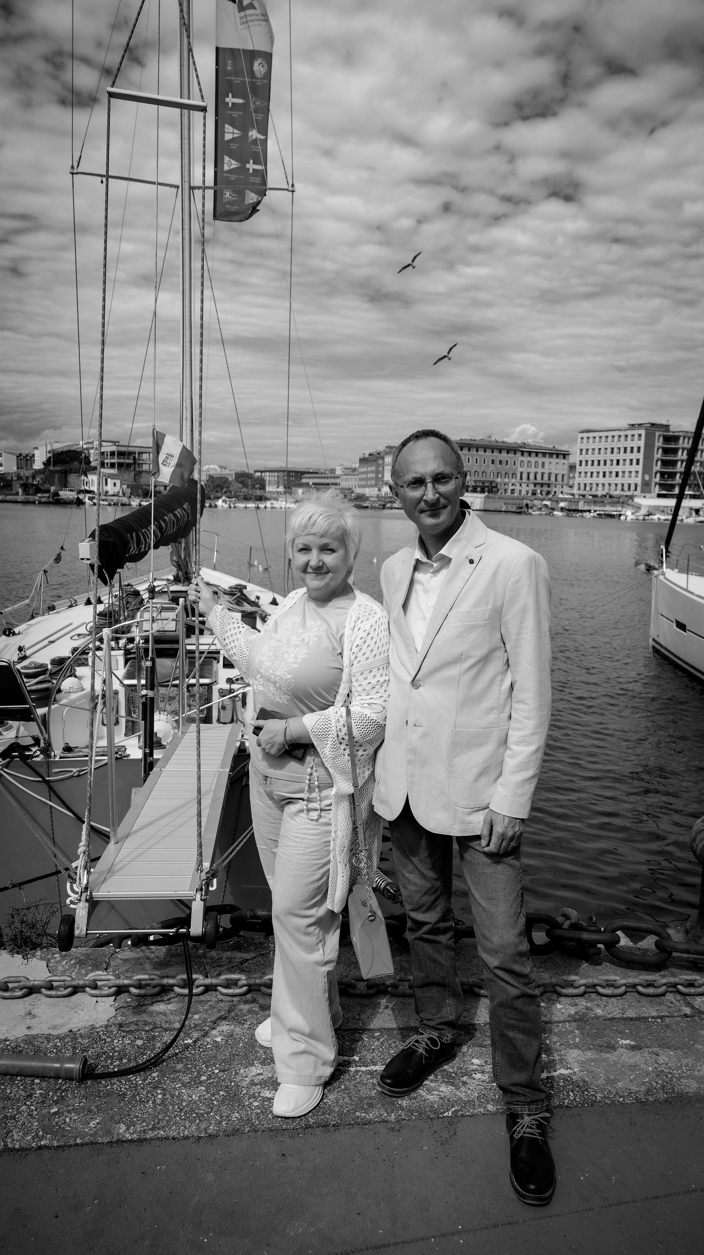 Черно-белая фотография пары, позирующей на причале в гавани, стоящей перед парусной лодкой и видом на городской пейзаж под облачным небом с летающими чайками.