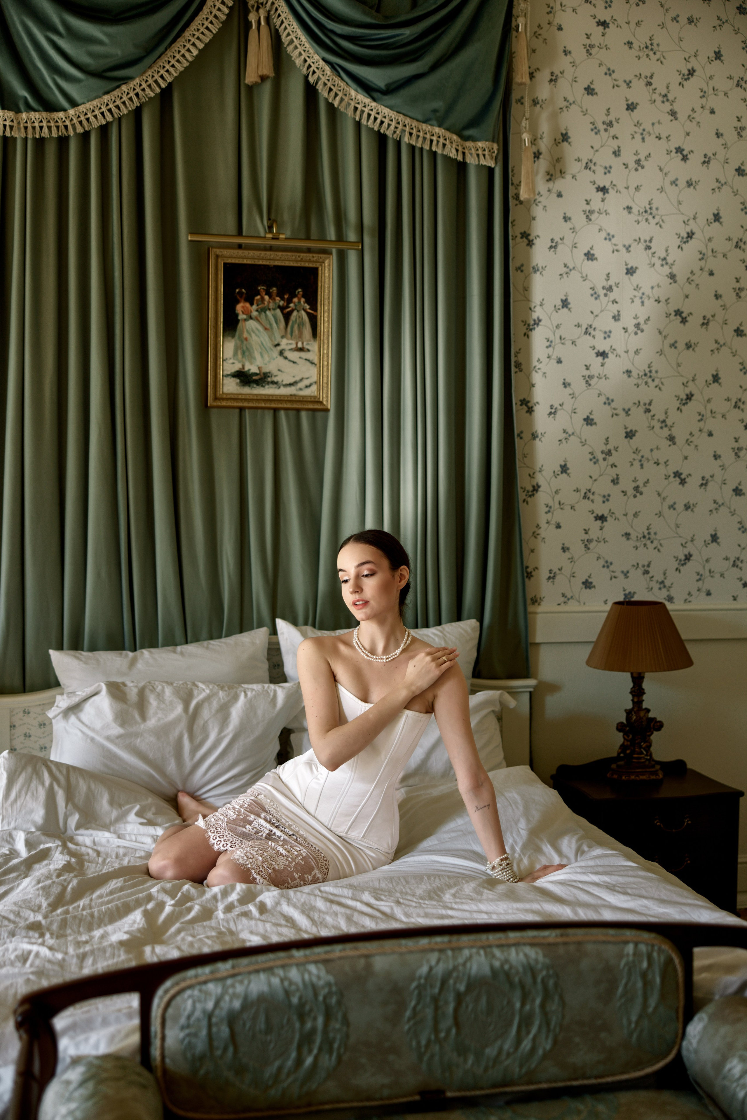 "Будуарная съемка в интерьере — модель полулежит на кровати, опираясь на локти, простыни небрежно смяты, приглушенный свет."