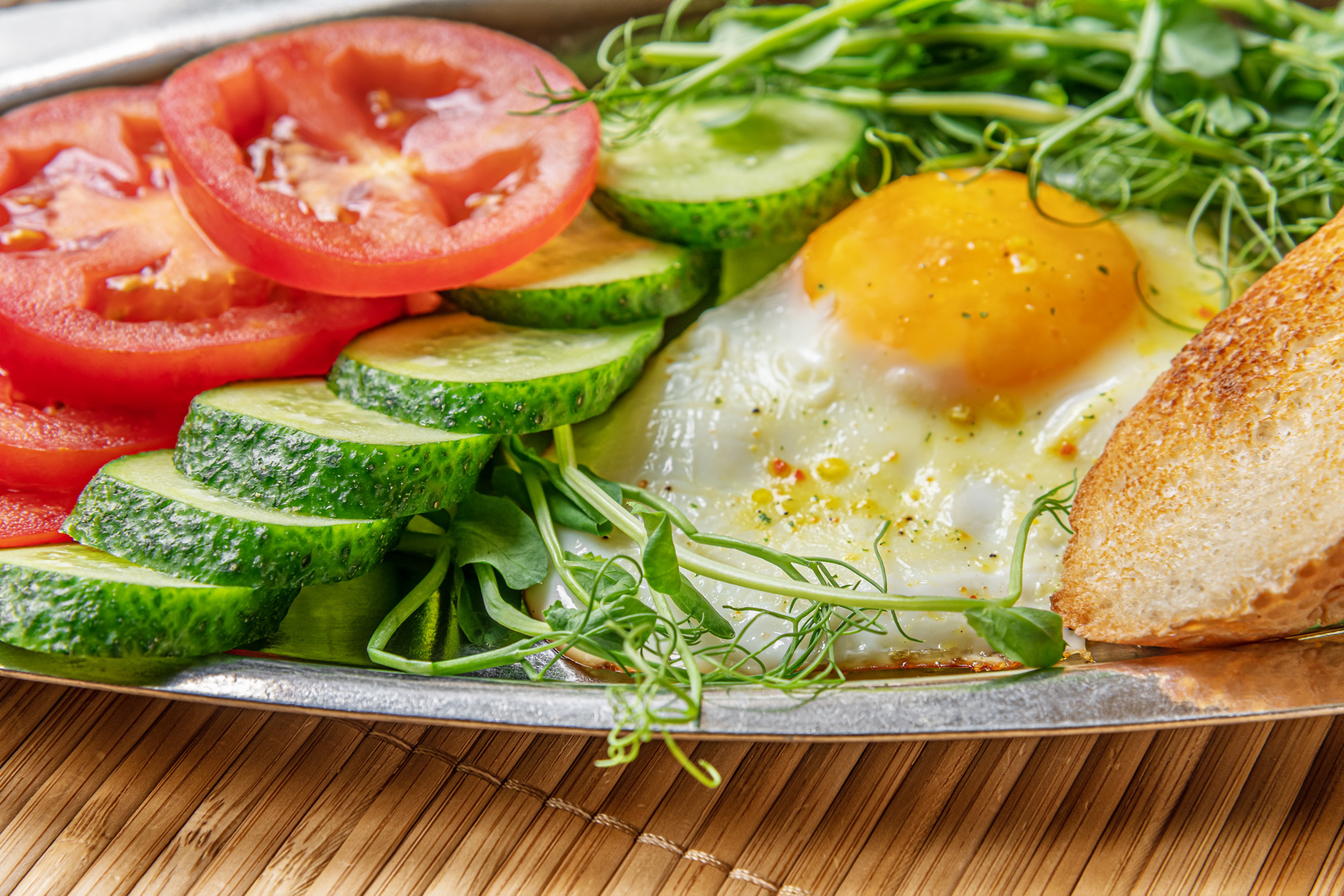 fried egg, toasted bread, cucumber and tomato slices and microgreens on a metal plate on bamboo straw surface, healthy breakfast concept
