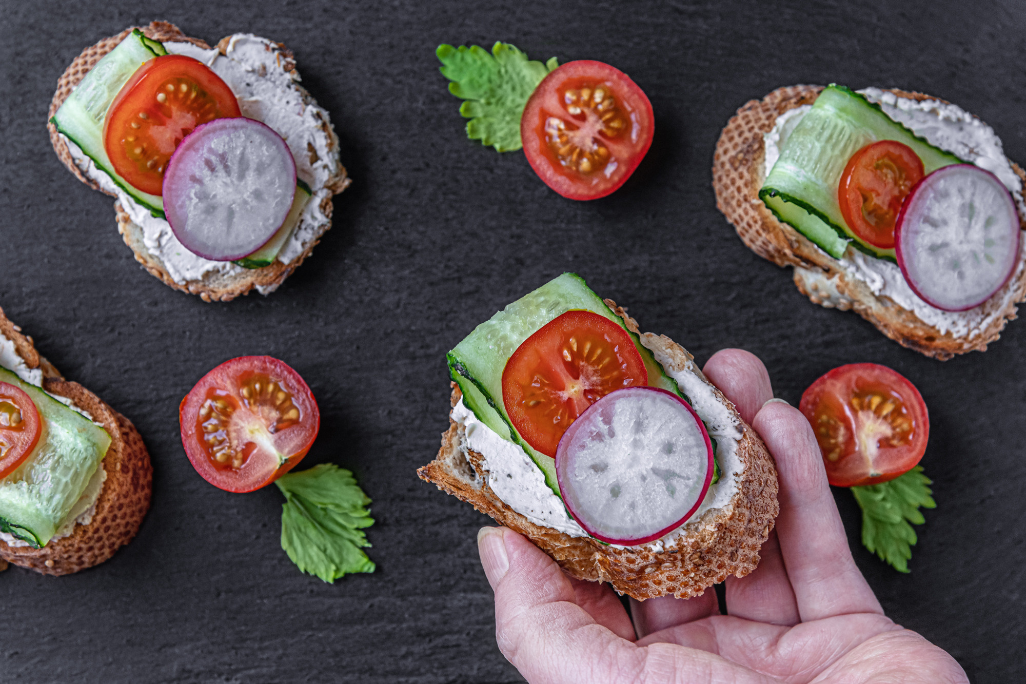 toasted baguette slices with cream cheese, cucumber, radish and tomato on a black slate board and female hand taking one