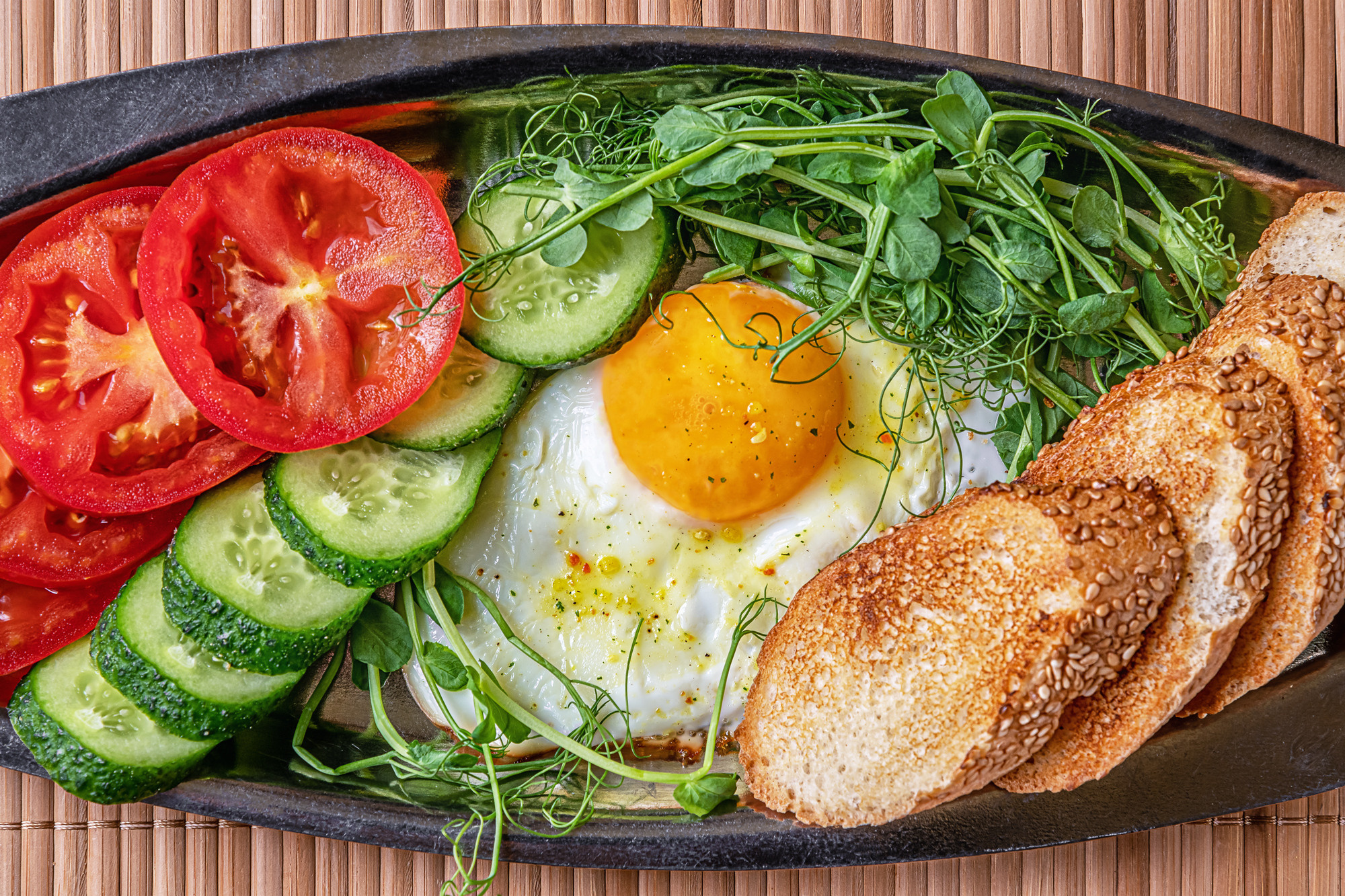 fried egg, toasted bread, cucumber and tomato slices and microgreens on a metal plate top view