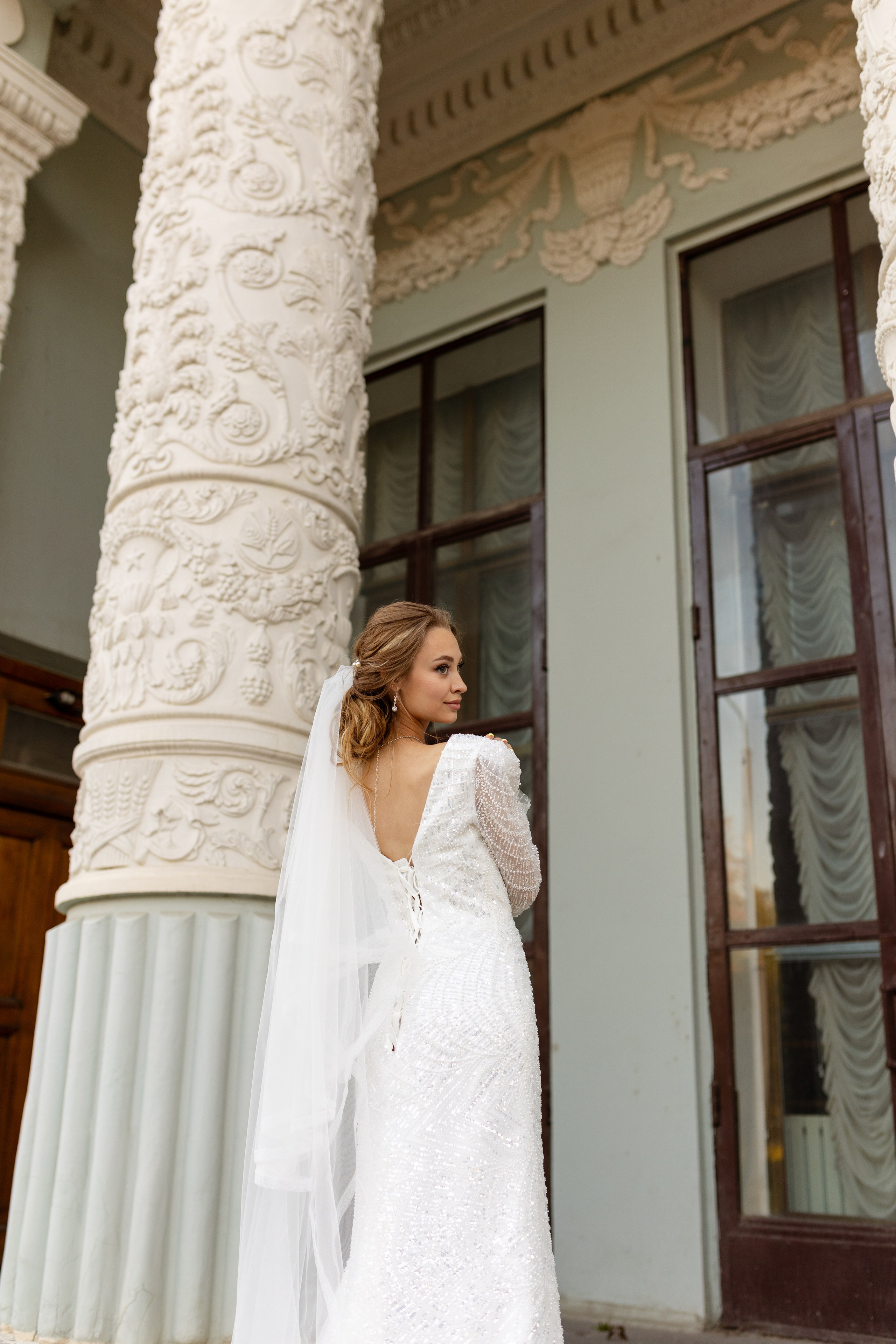 Невеста стоит у колонны, сбоку струится вуаль.