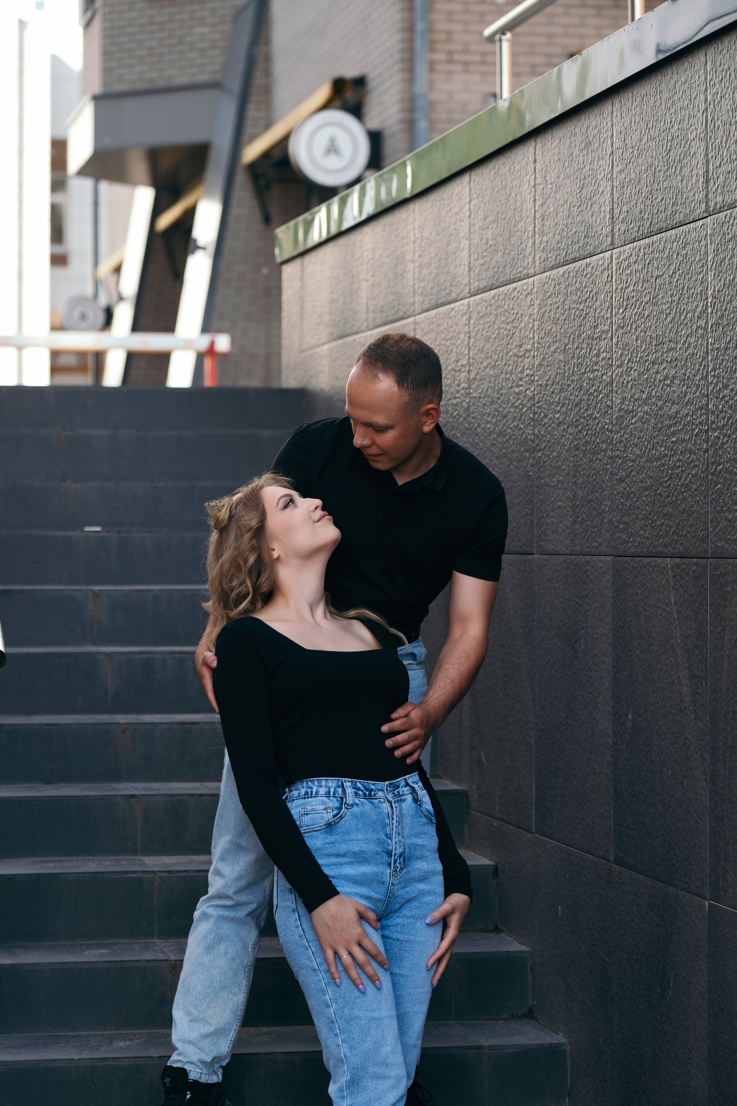 Молодая пара мужчина и девушка обнимаются городских локациях в черных футболках и синих джинсах стоя на серой лестнице, лав стори для влюбленной пары на кануне свадьбы