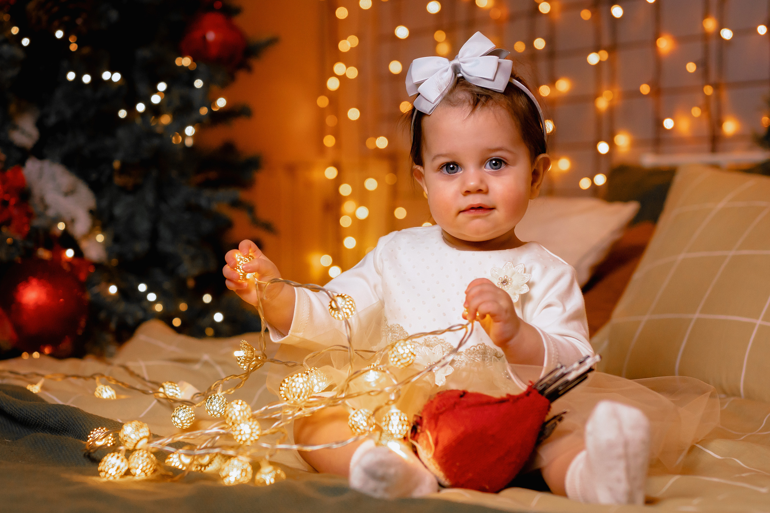 Годовалая девочка с большими глазами в белом платье и с бантом на голове сидит на кровати с гирляндой в руках на фоне новогодней ёлки