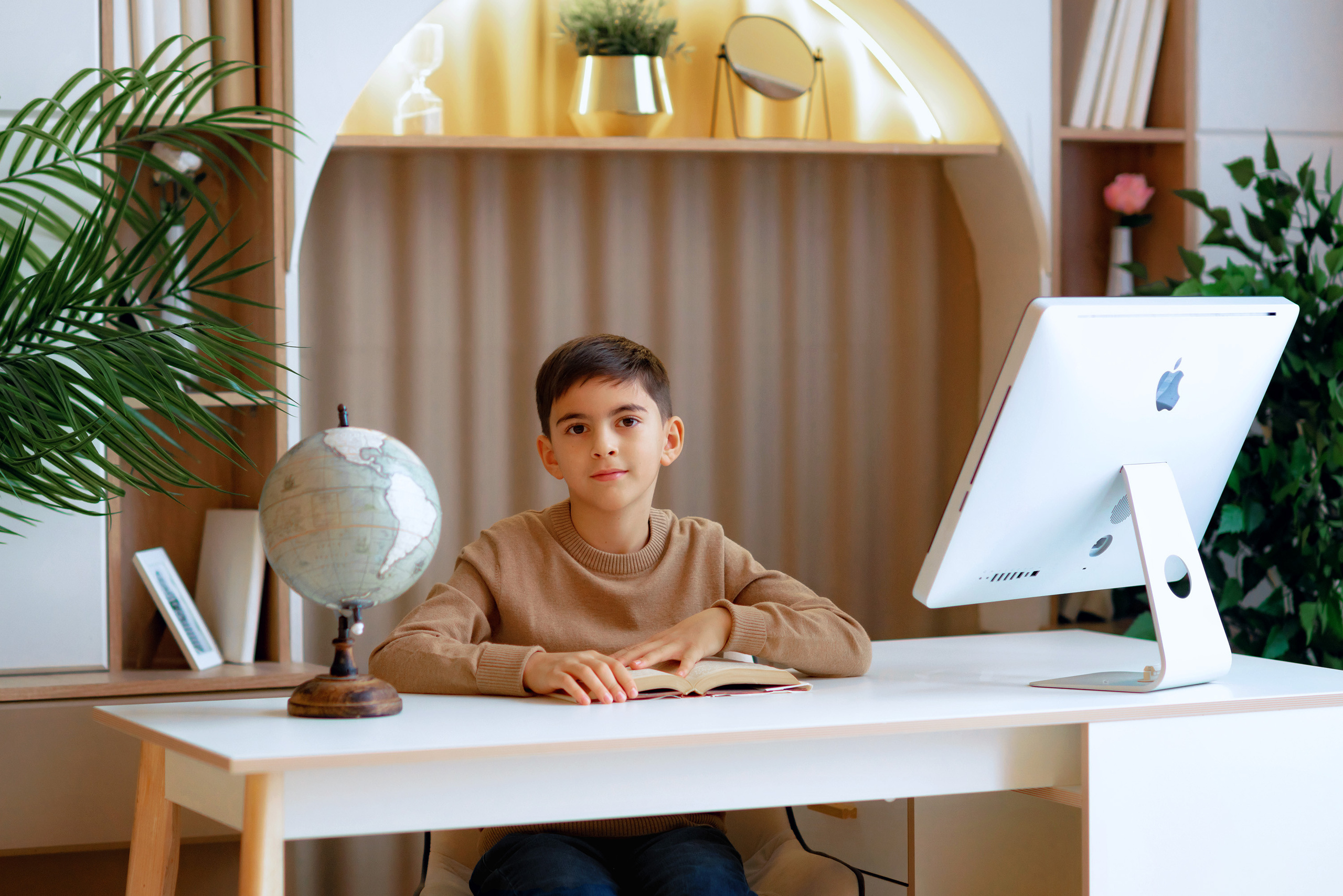 Школьник сидит за рабочим столом с глобусом, компьютером и книгой