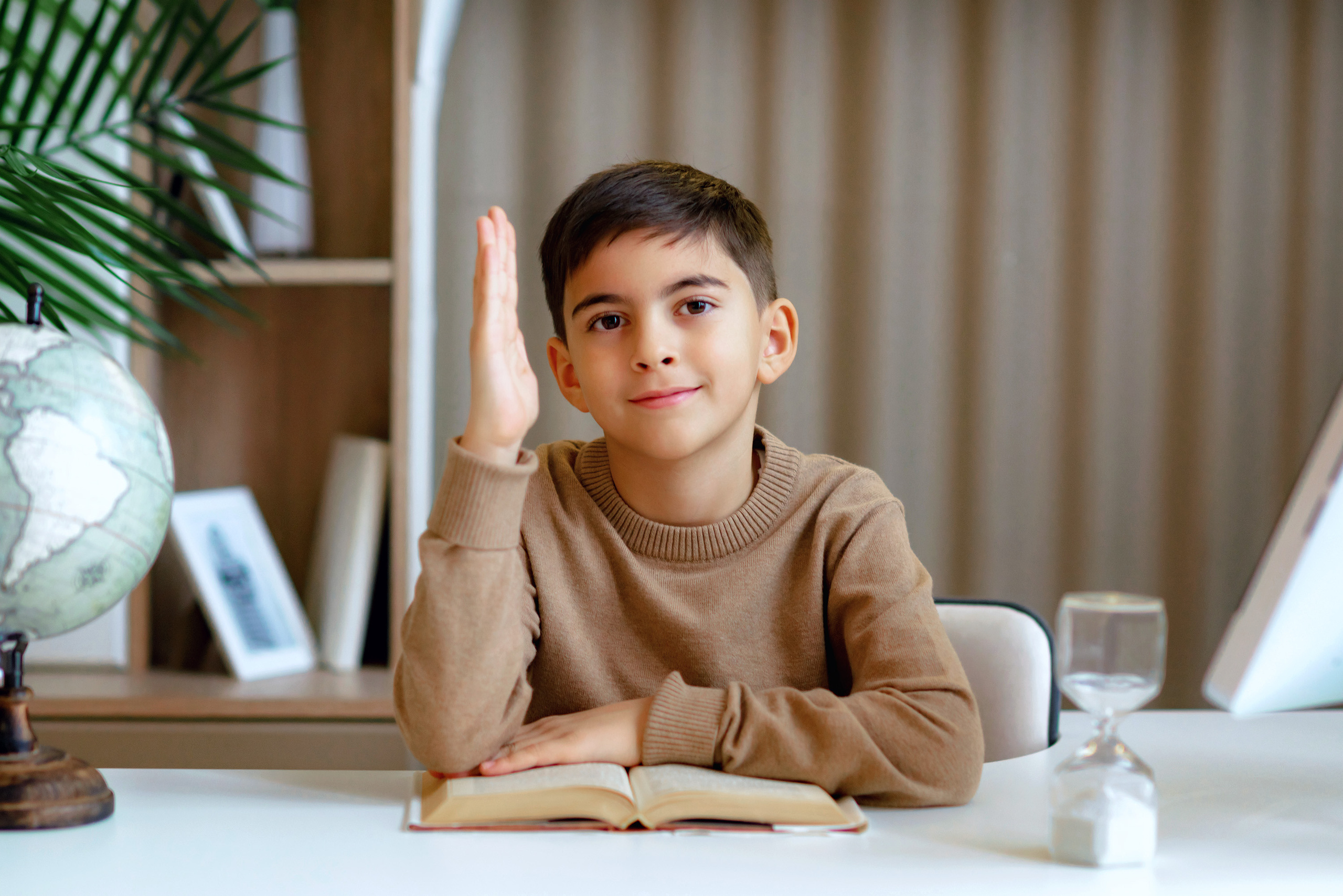 Школьник тянет руку за партой с песочными часами, глобусом и книгой