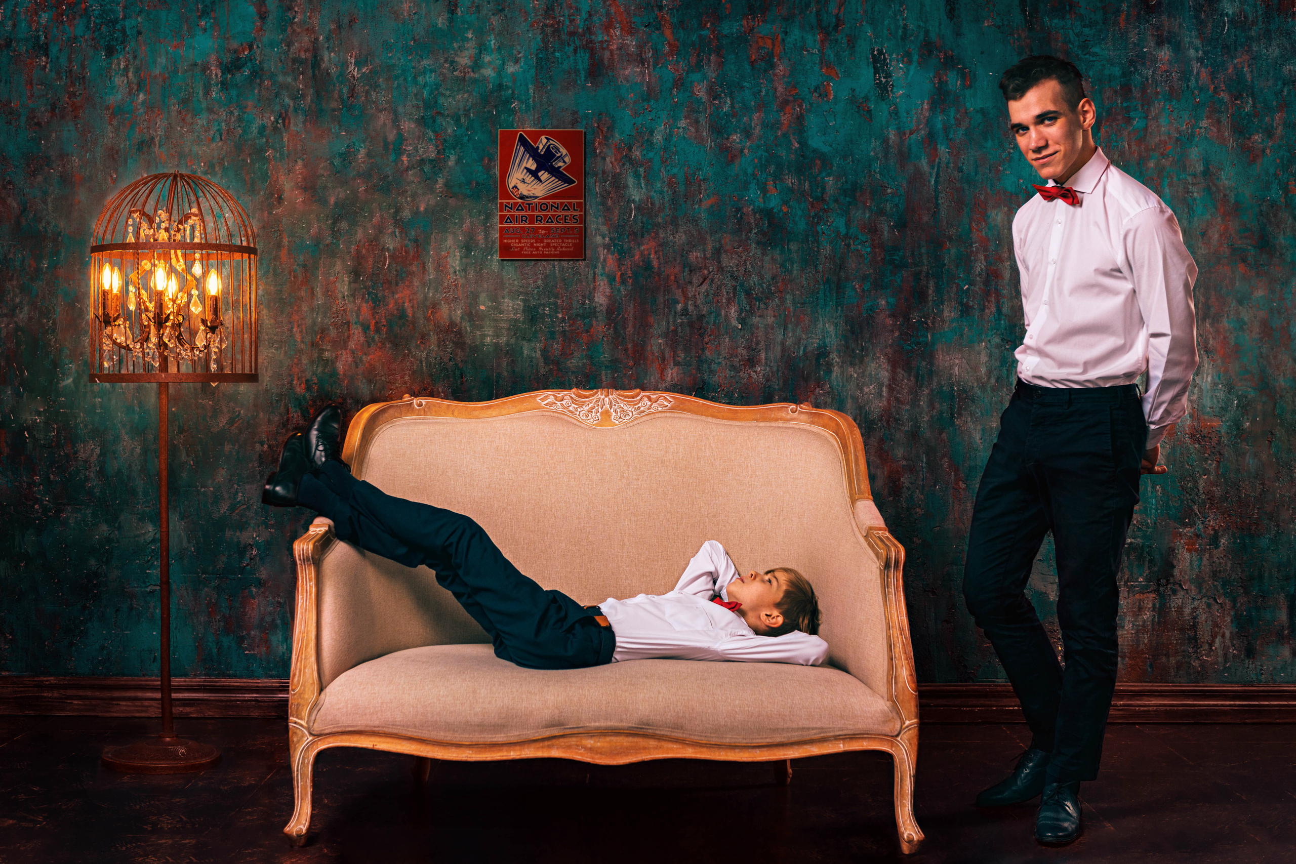 Два брата на фоне зеленой стены и лампы, один стоит, второй лежит на кресле