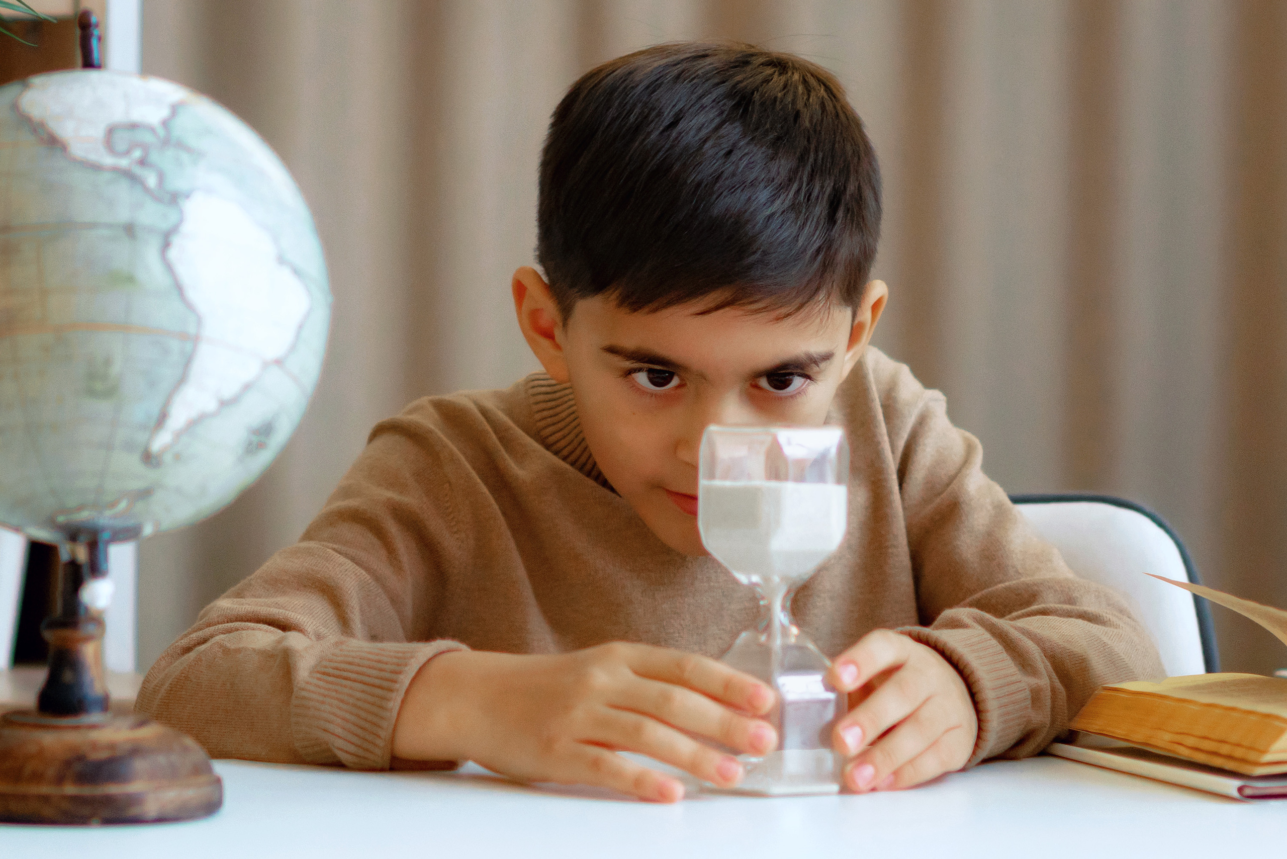 Школьник рассматривает песочные часы за партой с глобусом и книгой