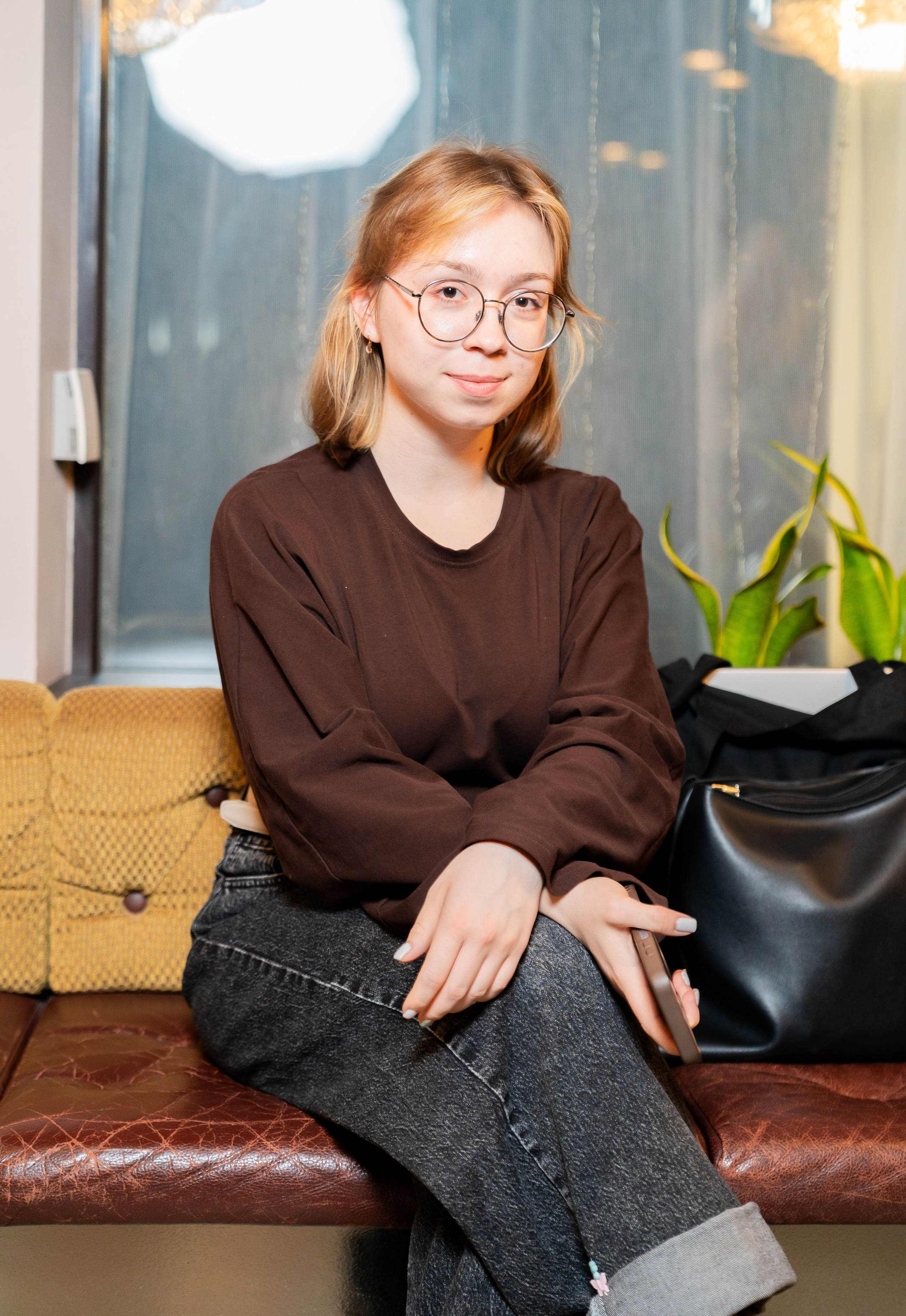 Портрет молодой девушки в очках в коричневом свитшоте на кожаном диване во время мероприятия в Radisson Sonya. Фотограф Евгений Мальцев, https://evrednix.ru.