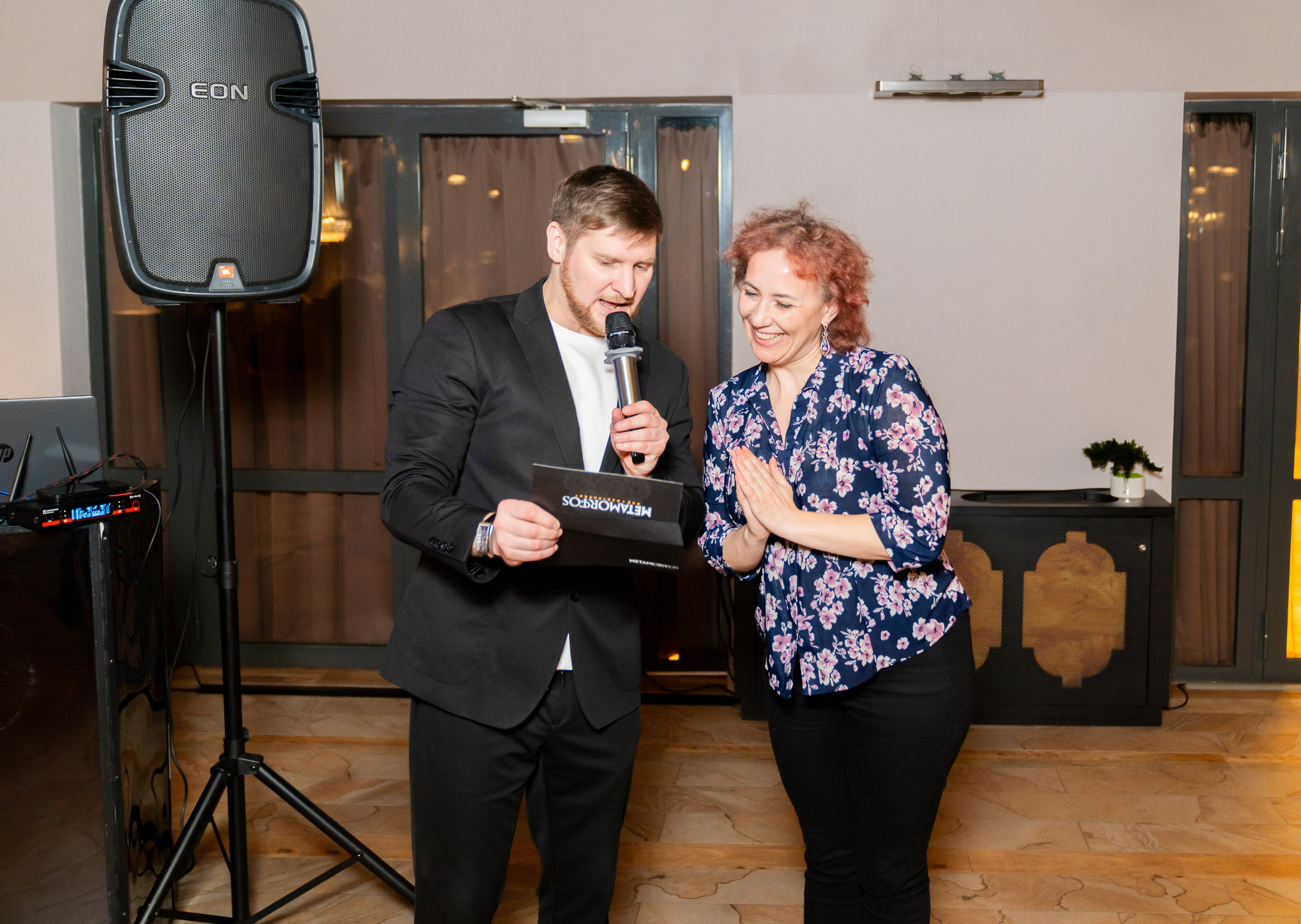 Вручение подарка: мужчина в чёрном пиджаке с микрофоном вручает коробку METAMORPHOSIS женщине с рыжими волосами на вечере в Radisson Sonya. Фото Евгения Мальцева.