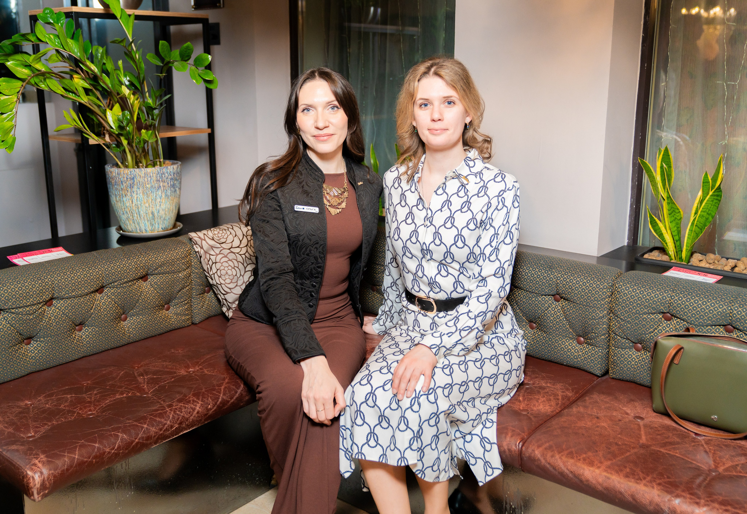 Две женщины на кожаном диване в лобби Radisson Sonya: одна в чёрном жакете, вторая в белом платье с синим принтом. Фото Евгения Мальцева.