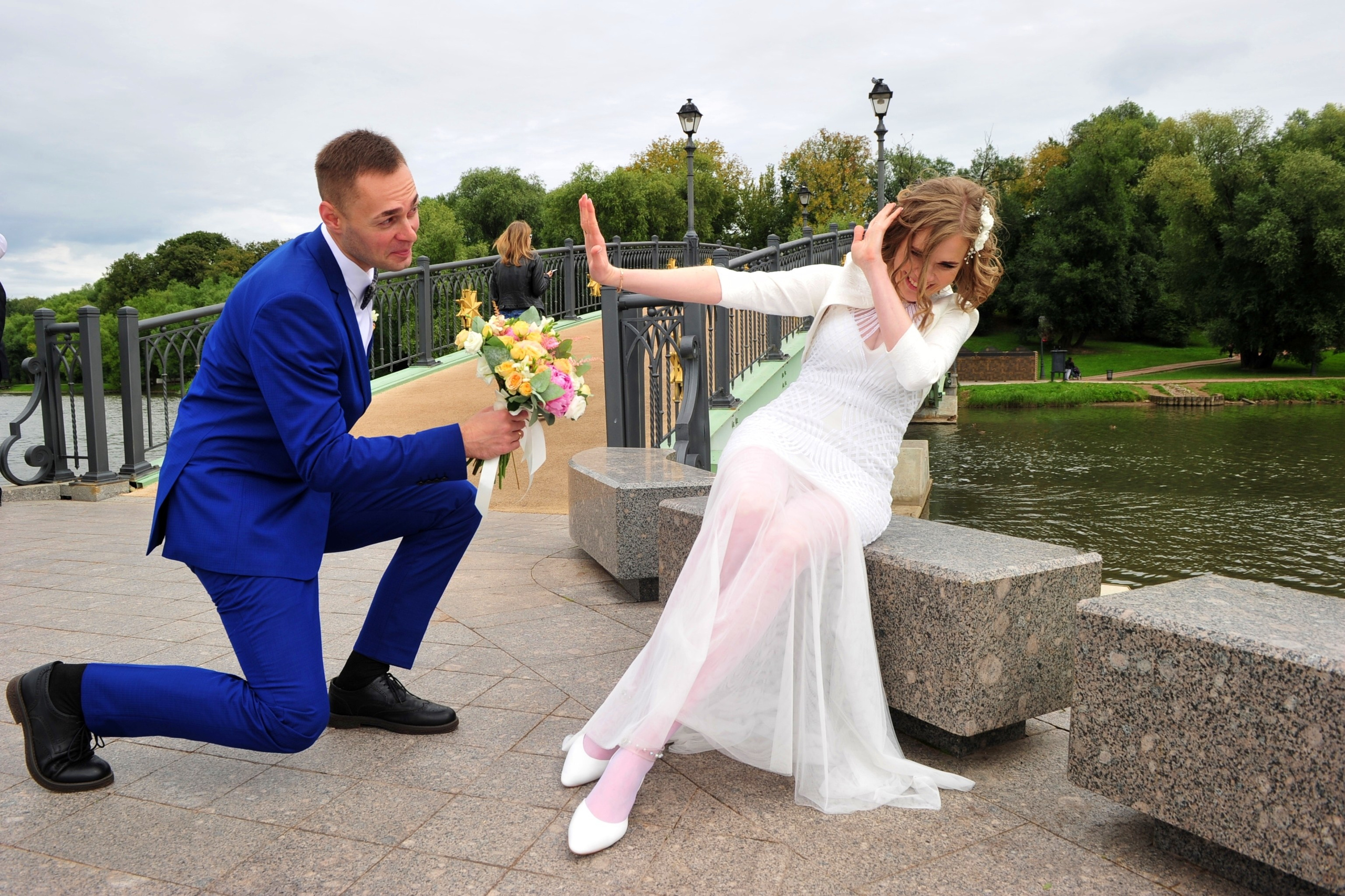 Свадьба в Царицыно хмурым прохладным летним днём, жених стоит на одном колене перед невестой и дарит ей букет, невеста отвергает букет от жениха, молодожены на фоне пруда у моста