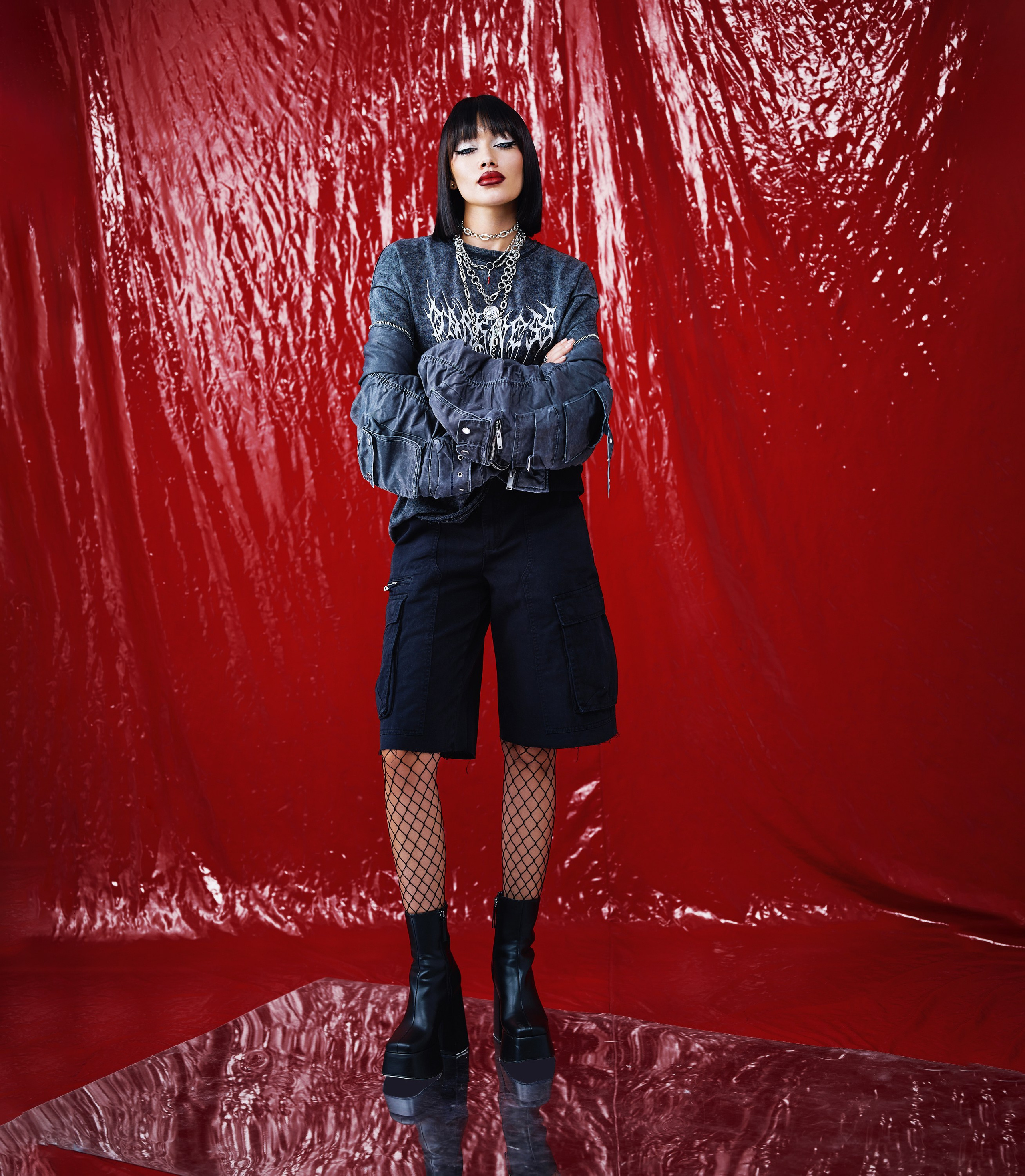 Полноразмерное фото модели в черной одежде с серебряными украшениями, на фоне ярко-красной стены, модель стоит со скрещенными руками и уверенной позой
