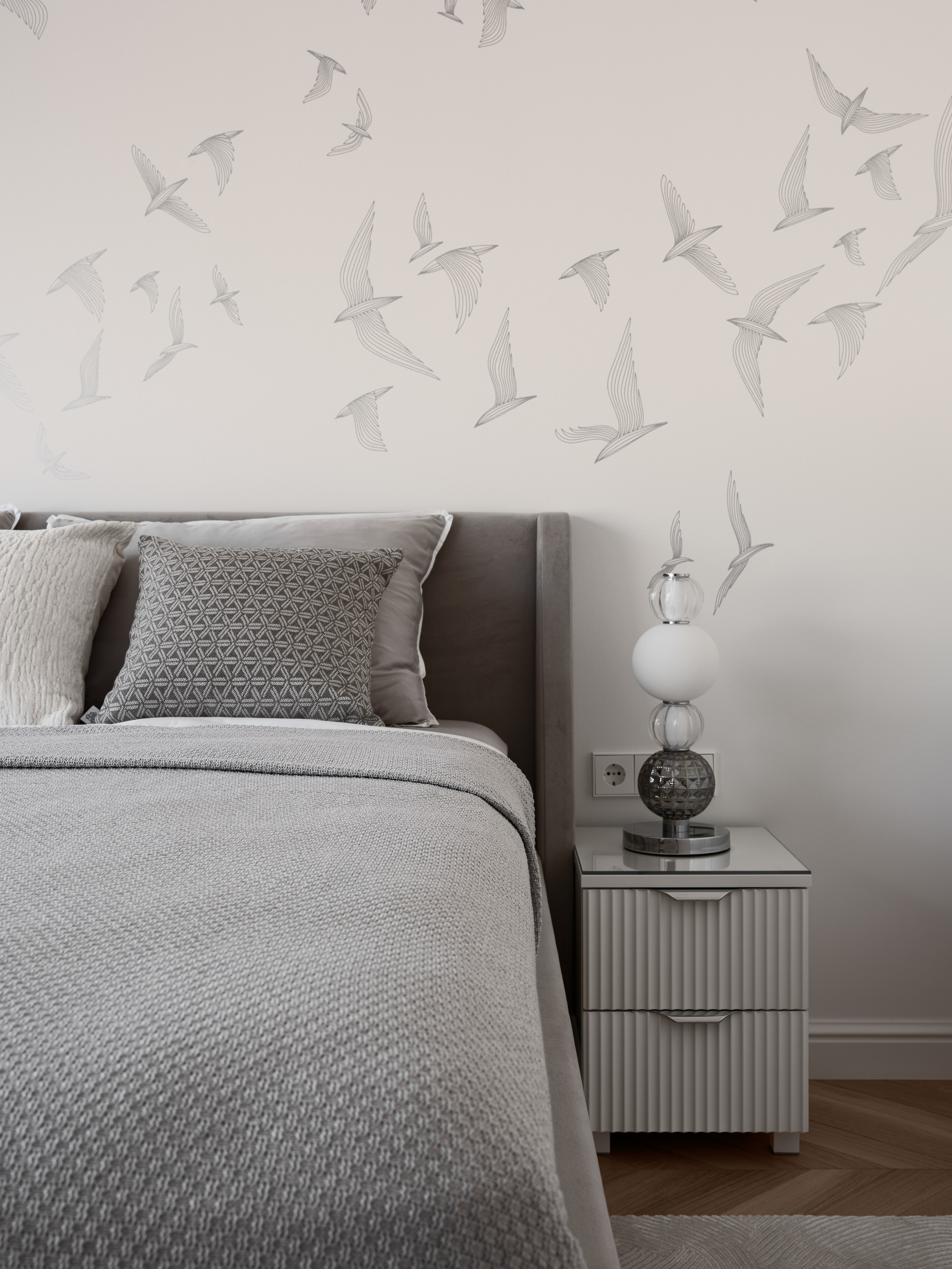 Современный интерьер спальни с серым текстилем, белыми подушками и прикроватной тумбой, оформленный дизайнером Алёной Боярской. Наталья Якушева создала сдержанную композицию с лампой и графикой птиц, а Сергей Денисенко передал воздушность и уют пространства.