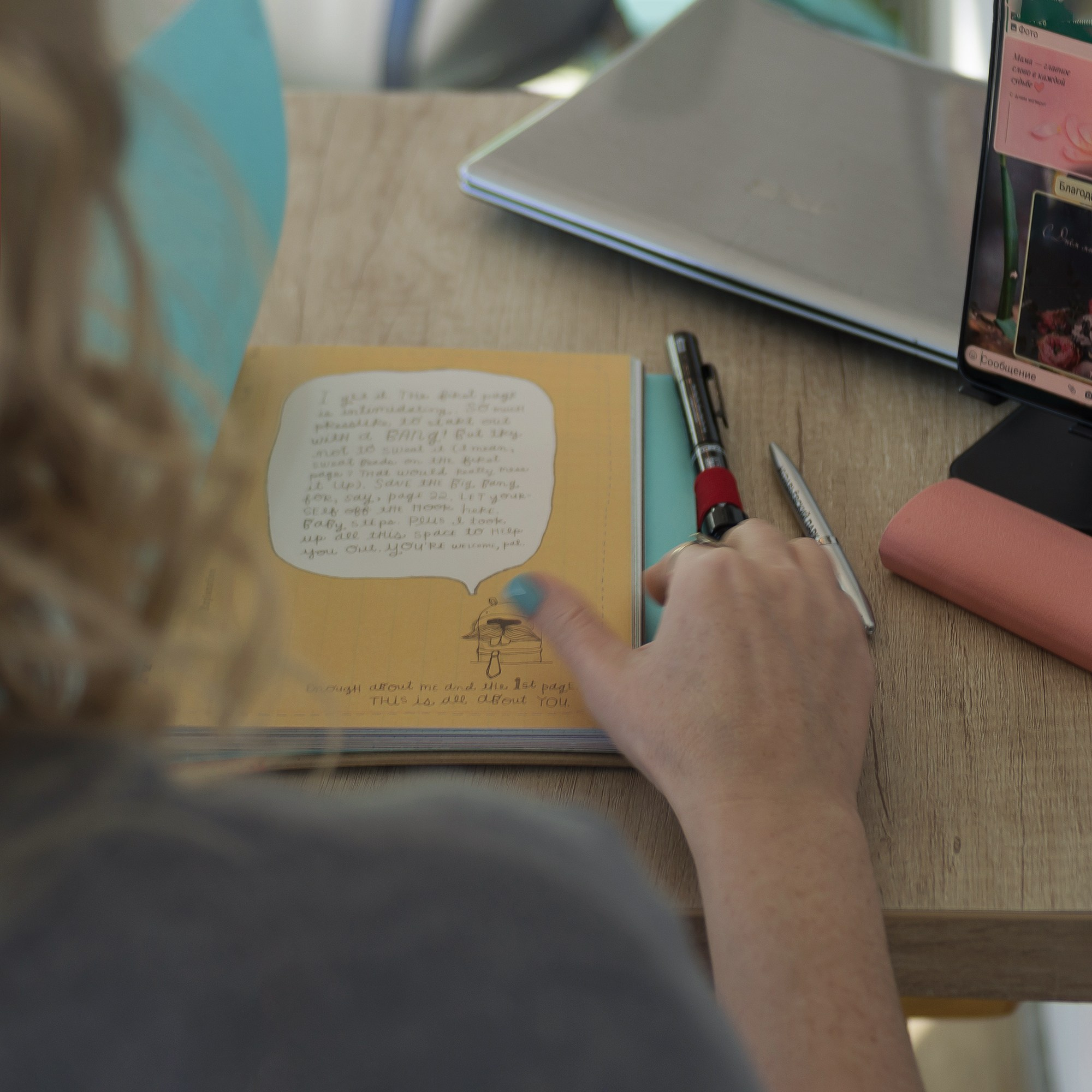 Взгляд из-за спины на рабочий стол. Ноутбук, записная книжка, ручка.