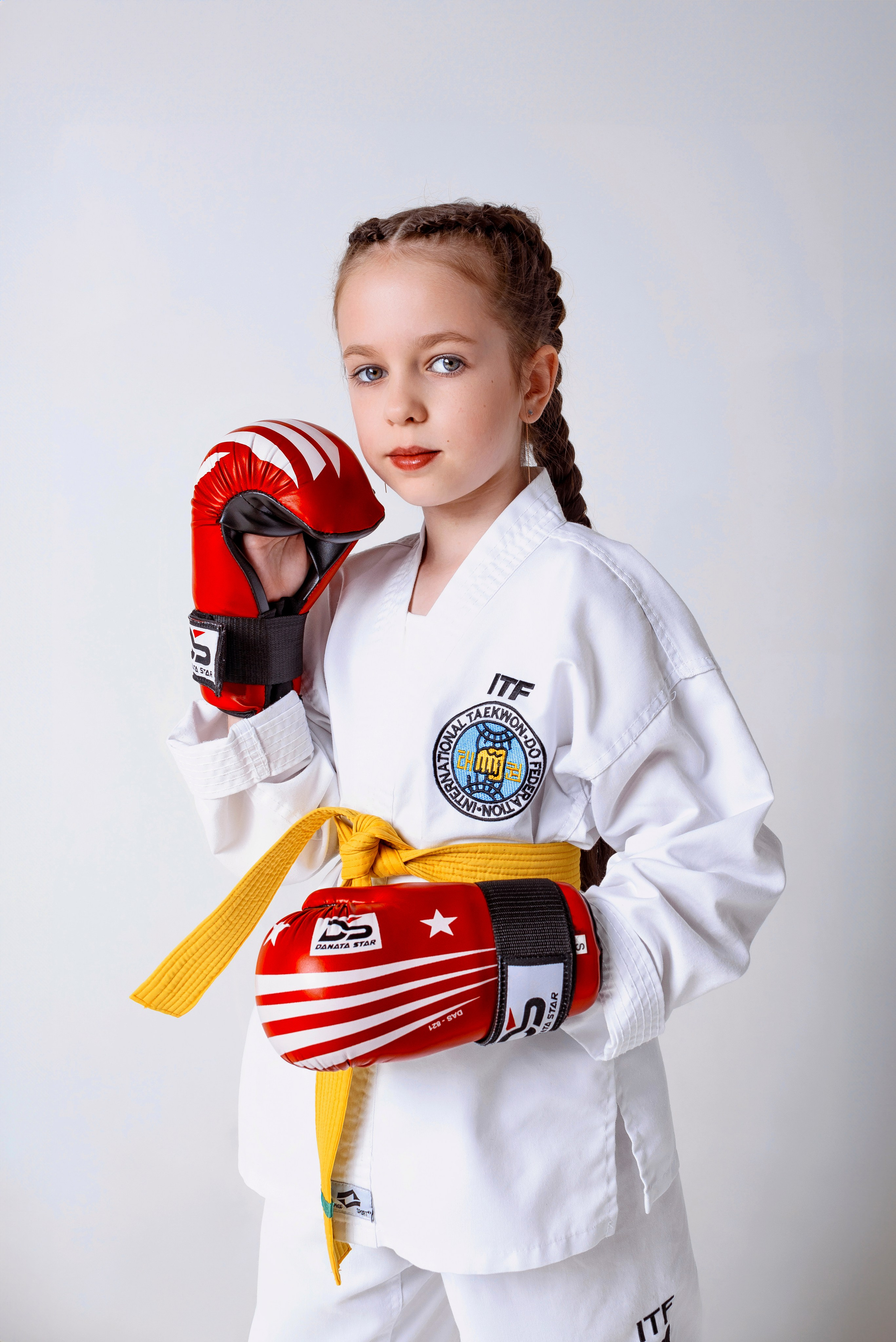 На изображении изображено лицо, одетое в белую форму Тхэквондо (добок) с желтым поясом. Человек носит красные боксерские перчатки и, вероятно, готовится к тренировке боевых искусств. На форме есть нашивки, одна из которых содержит буквы "ITF", что, вероятно, указывает на Международную федерацию Тхэквондо. Фон светлый и однородный, что обеспечивает четкий обзор объекта.