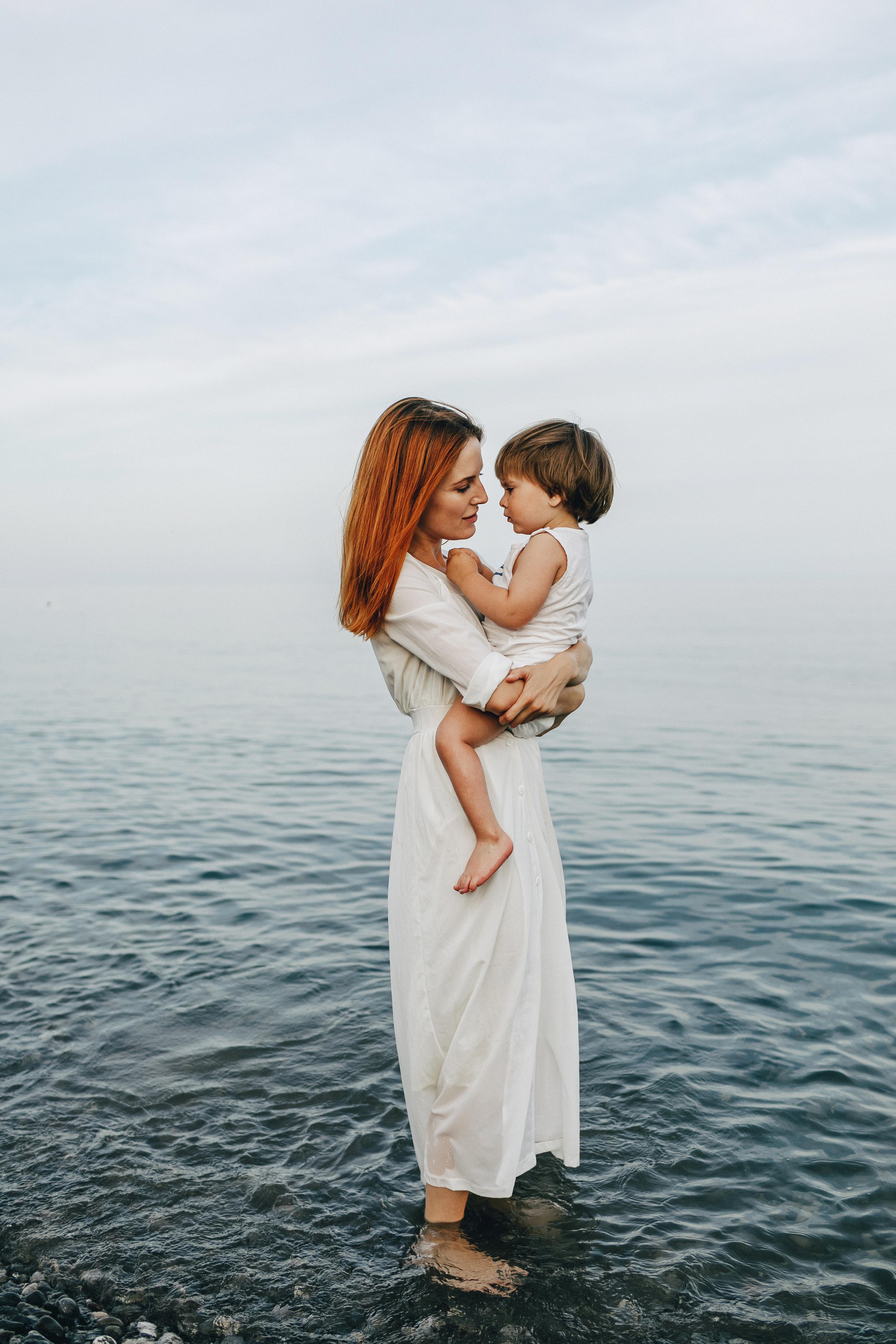 Мать держит своего ребенка на руках стоя босяком в море. 