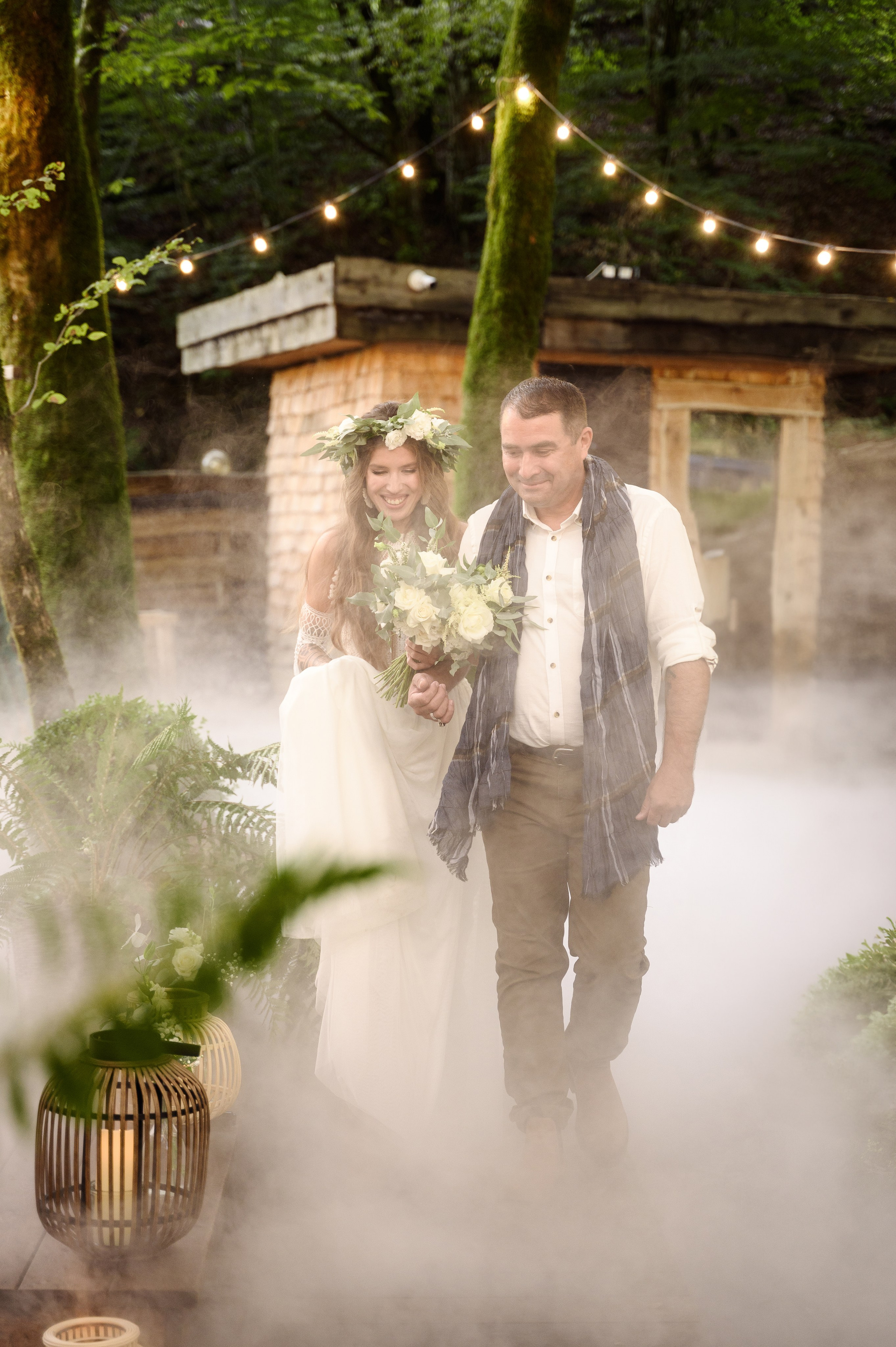 Очень нежный кадр где отец ведёт дочь на церемонии свадебной к её будущему мужу это происходит в лесу сверху натянуты гирлянды и всё заполнено белым туманом