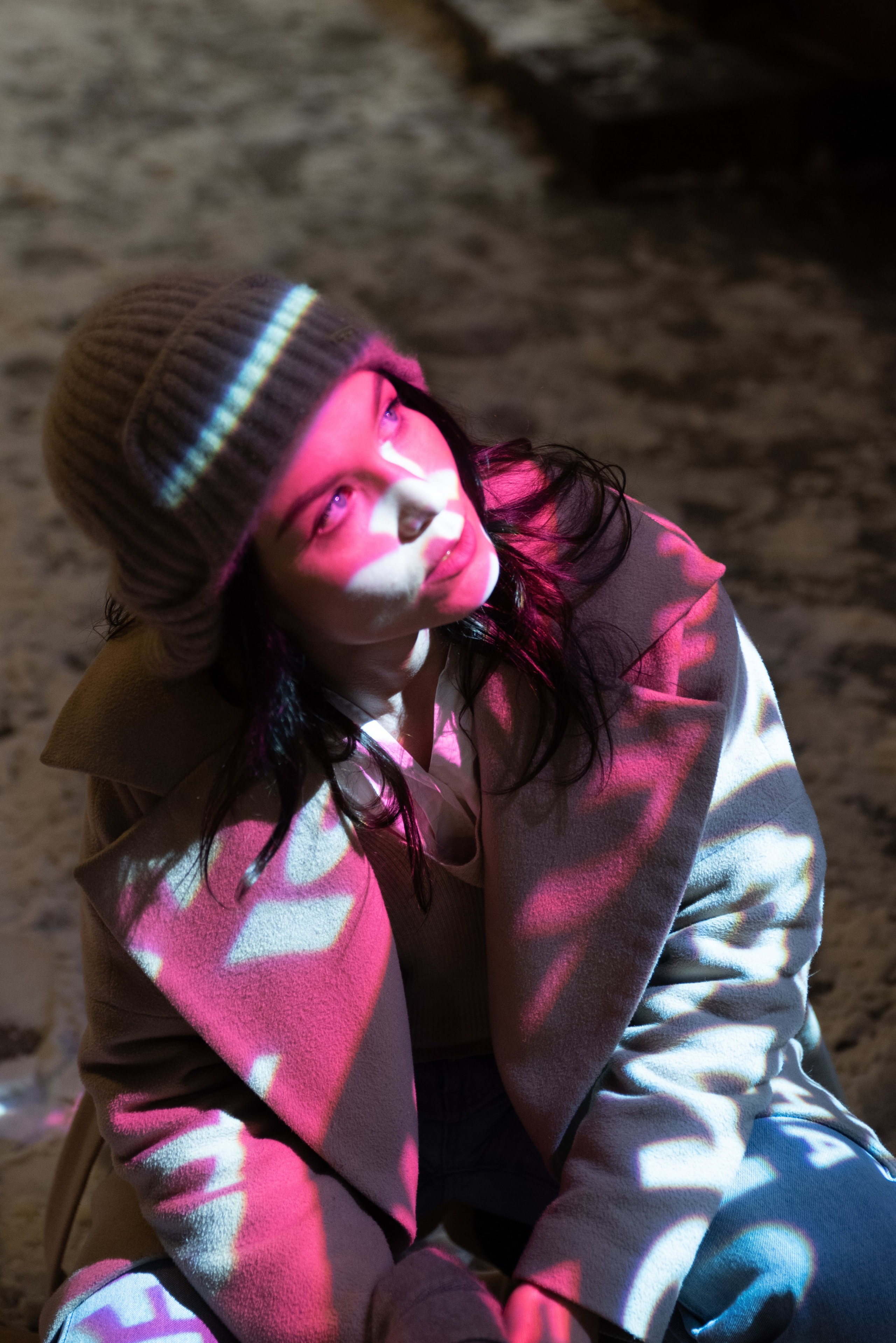 Зима, снег, улица, молодая женщина в пальто и вязаной шапке сидит на корточках, на лице и на одежде красные блики от рекламы